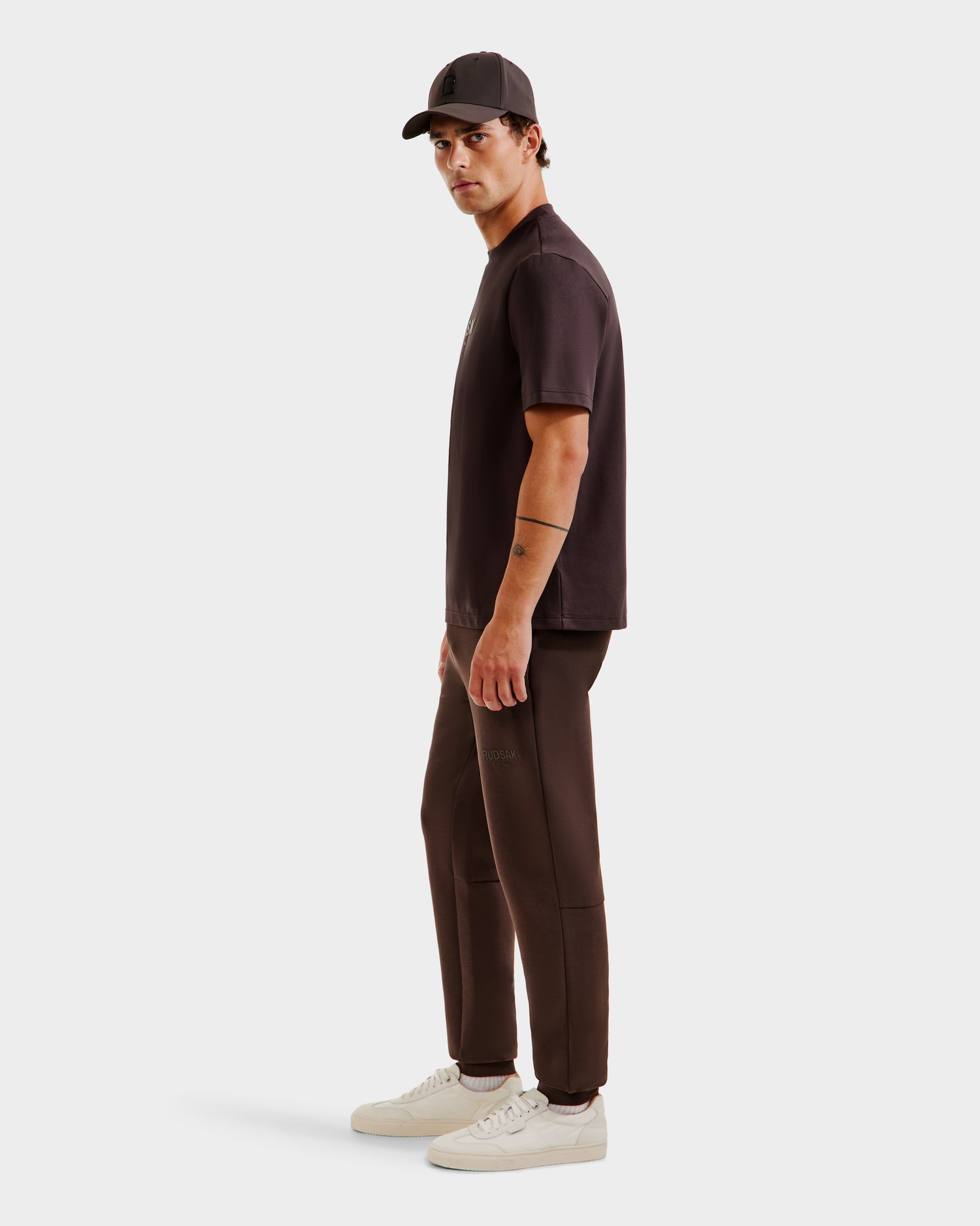 A man stands sideways in a Rudsak CASEY - 6525037 Espresso mens T-shirt and matching joggers, paired with a black cap and white sneakers—a comfortable essential look set against a plain light background.