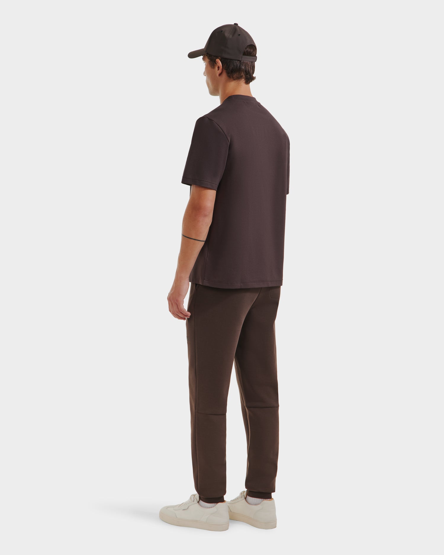 A person stands facing away, wearing the Rudsak CASEY - 6525037 Espresso men’s T-shirt, paired with a dark cap, dark pants, and white sneakers against a plain white background.