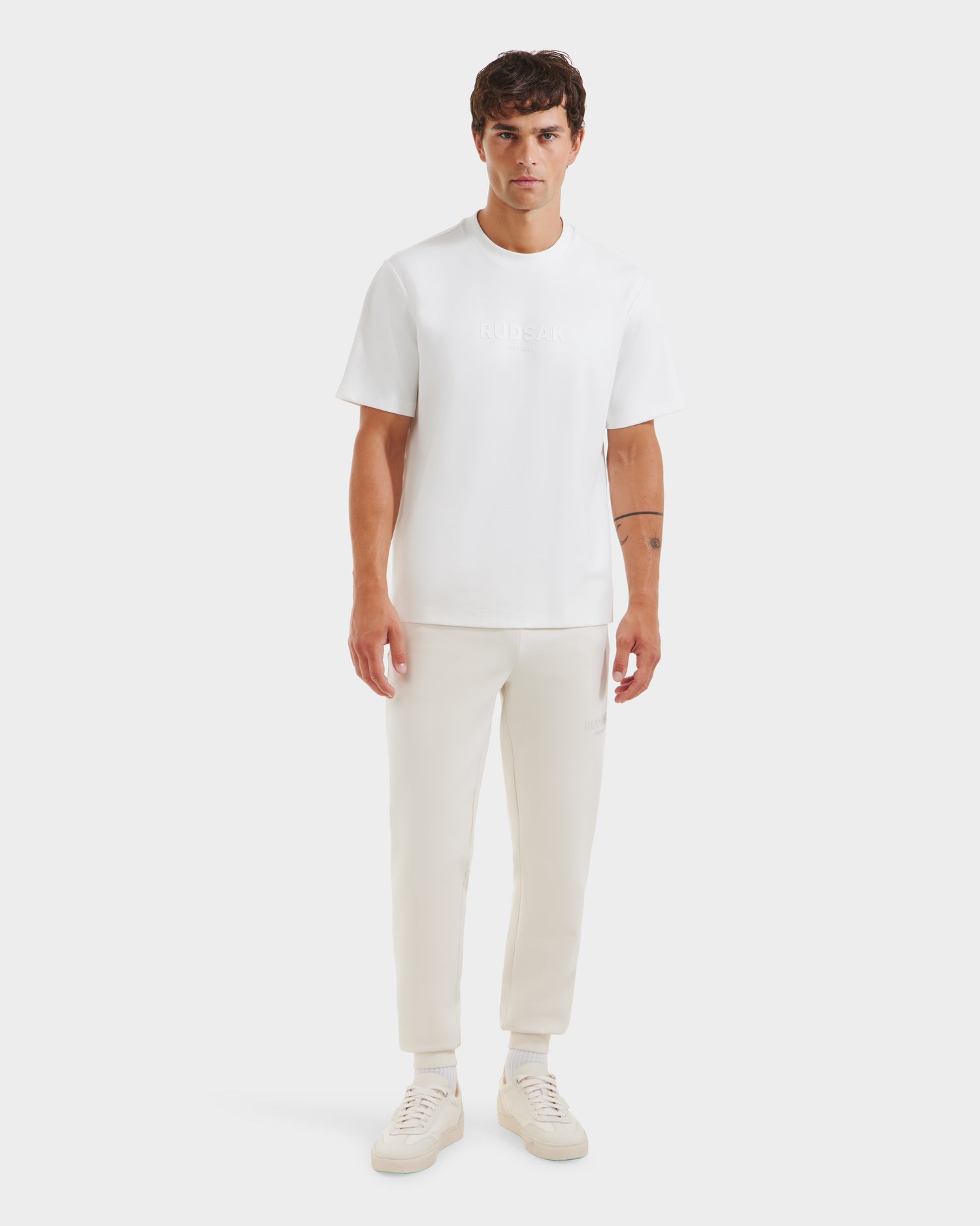 A man faces forward wearing the Rudsak CASEY - 6525037 Optic White mens T-shirt, paired with white pants and sneakers, highlighting this comfortable essential against a plain white background.