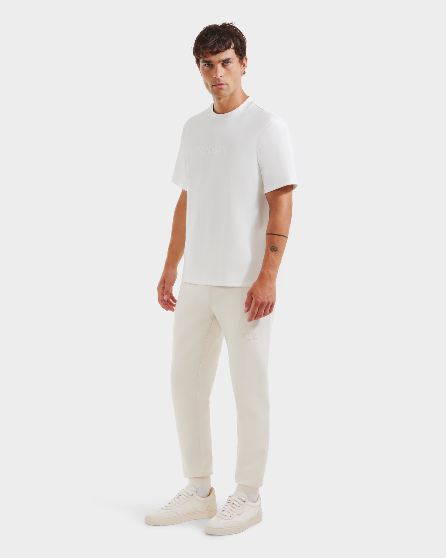 A man with short dark hair and a neutral expression stands against a white background, wearing the Rudsak CASEY - 6525037 Optic White essential men’s T-shirt, cream jogger pants, and white sneakers.