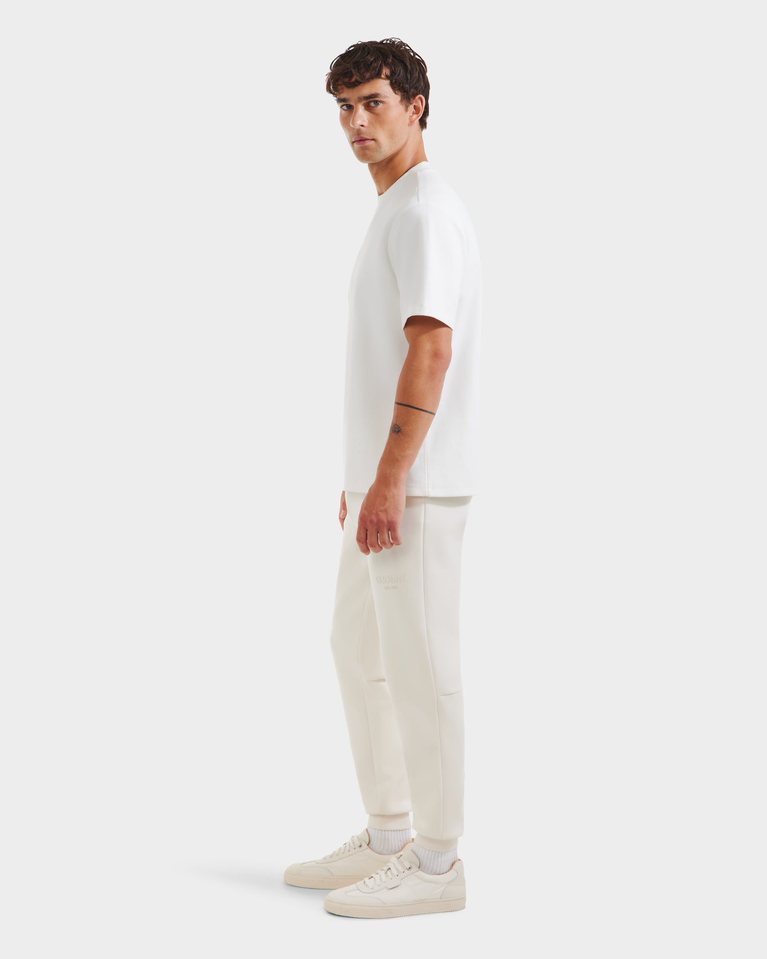 A man stands sideways facing left, wearing the Rudsak CASEY - 6525037 Optic White T-shirt, off-white jogger pants, and white sneakers. The comfortable outfit is set against a plain light gray background.