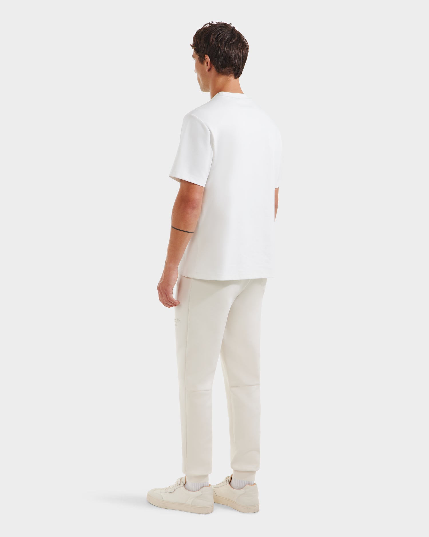 A man with short brown hair and a black armband tattoo on his left arm faces away, wearing the Rudsak CASEY - 6525037 Optic White T-shirt. Paired with white pants and sneakers, he stands against a plain, light background.