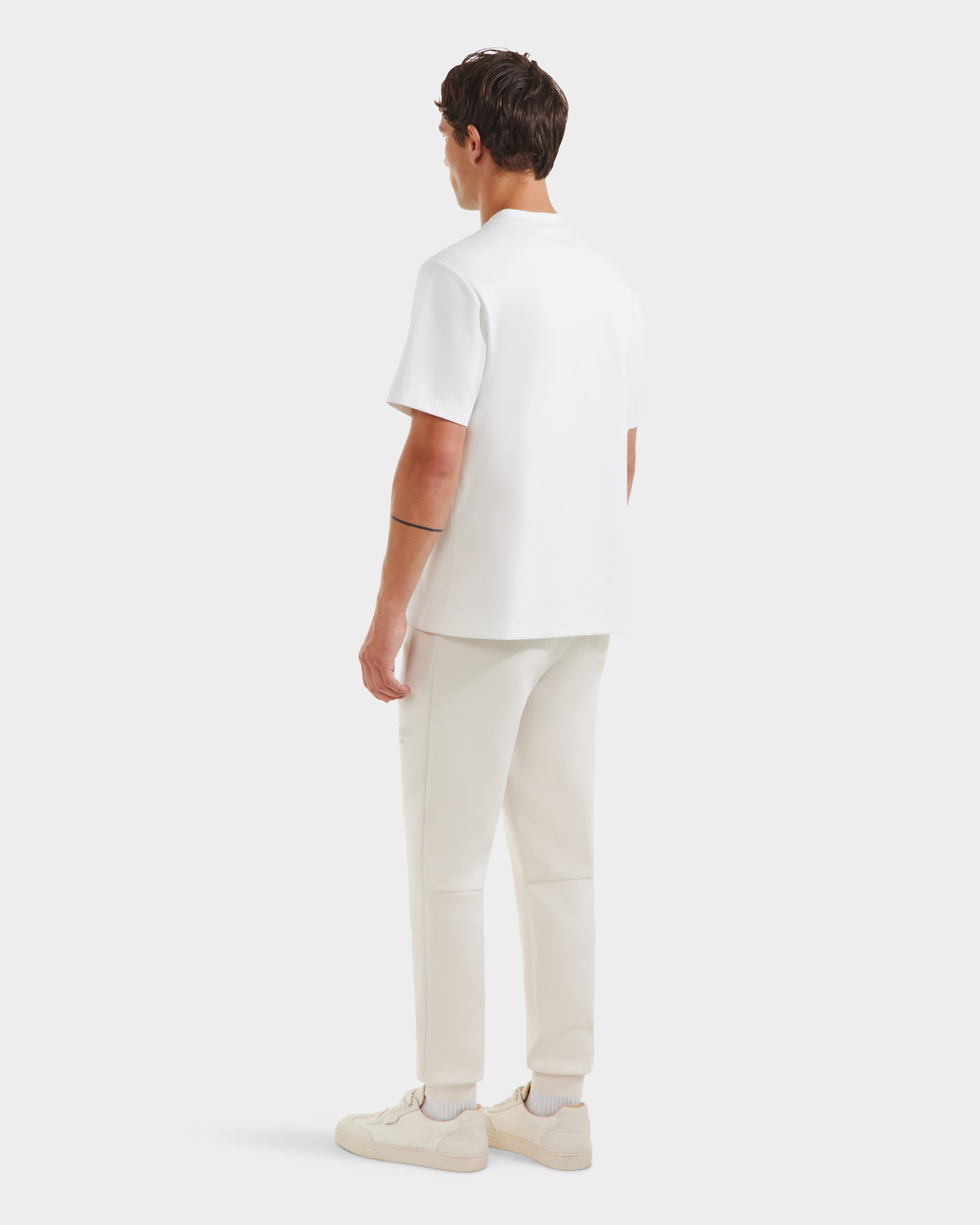 A man with short brown hair and a black armband tattoo on his left arm faces away, wearing the Rudsak CASEY - 6525037 Optic White T-shirt. Paired with white pants and sneakers, he stands against a plain, light background.