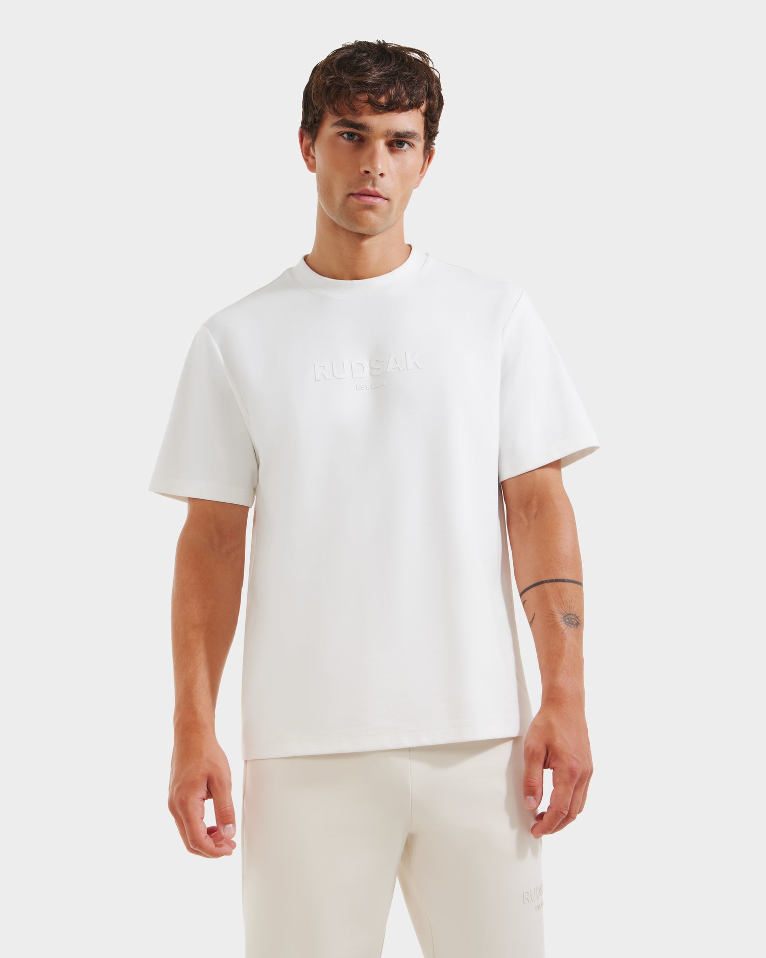 A man wears the Rudsak CASEY - 6525037 Optic White T-shirt, featuring subtle embossed chest text, paired with matching white pants against a light background. He has a simple tattoo on his left forearm.