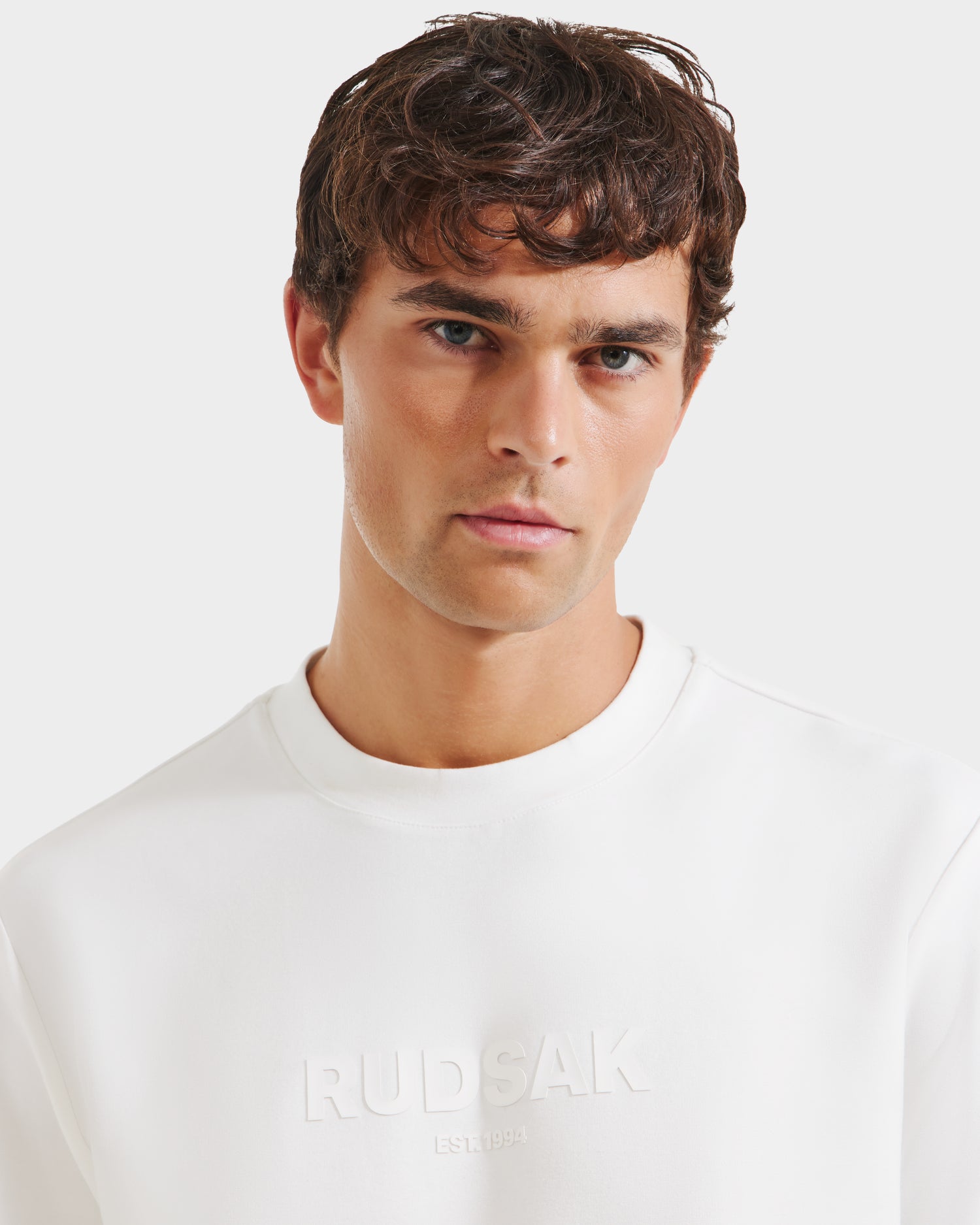 A young man with short, wavy brown hair wears the Rudsak CASEY - 6525037 sweatshirt in Optic White, standing against a white background and looking directly at the camera with a neutral expression.