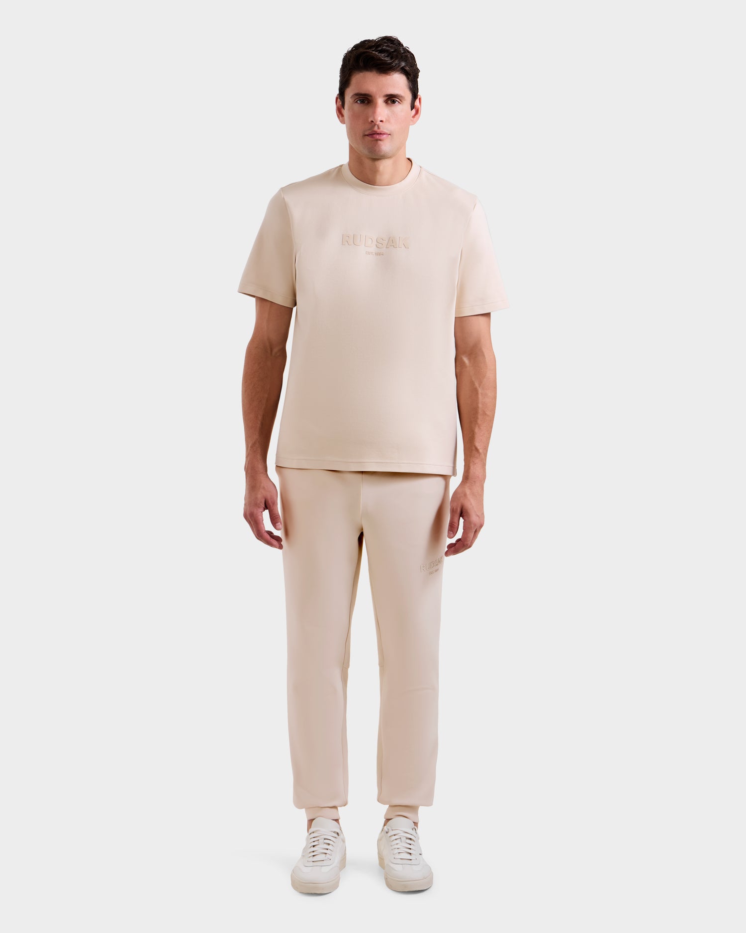 A man faces forward in the Rudsak CASEY - 6525037 Vanilla men’s T-shirt and matching pants set with white sneakers, standing against a plain white background.