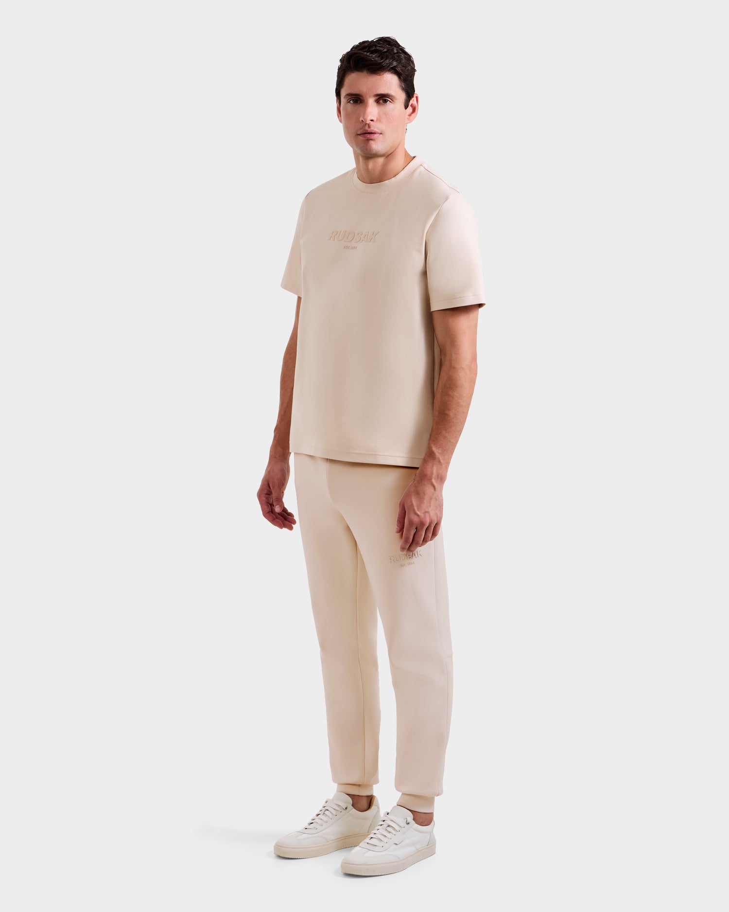 A man stands against a white background wearing the CASEY - 6525037 Vanilla T-shirt and jogger set from Rudsak, paired with white sneakers. He faces forward, relaxed, highlighting this stylish and comfortable essential.