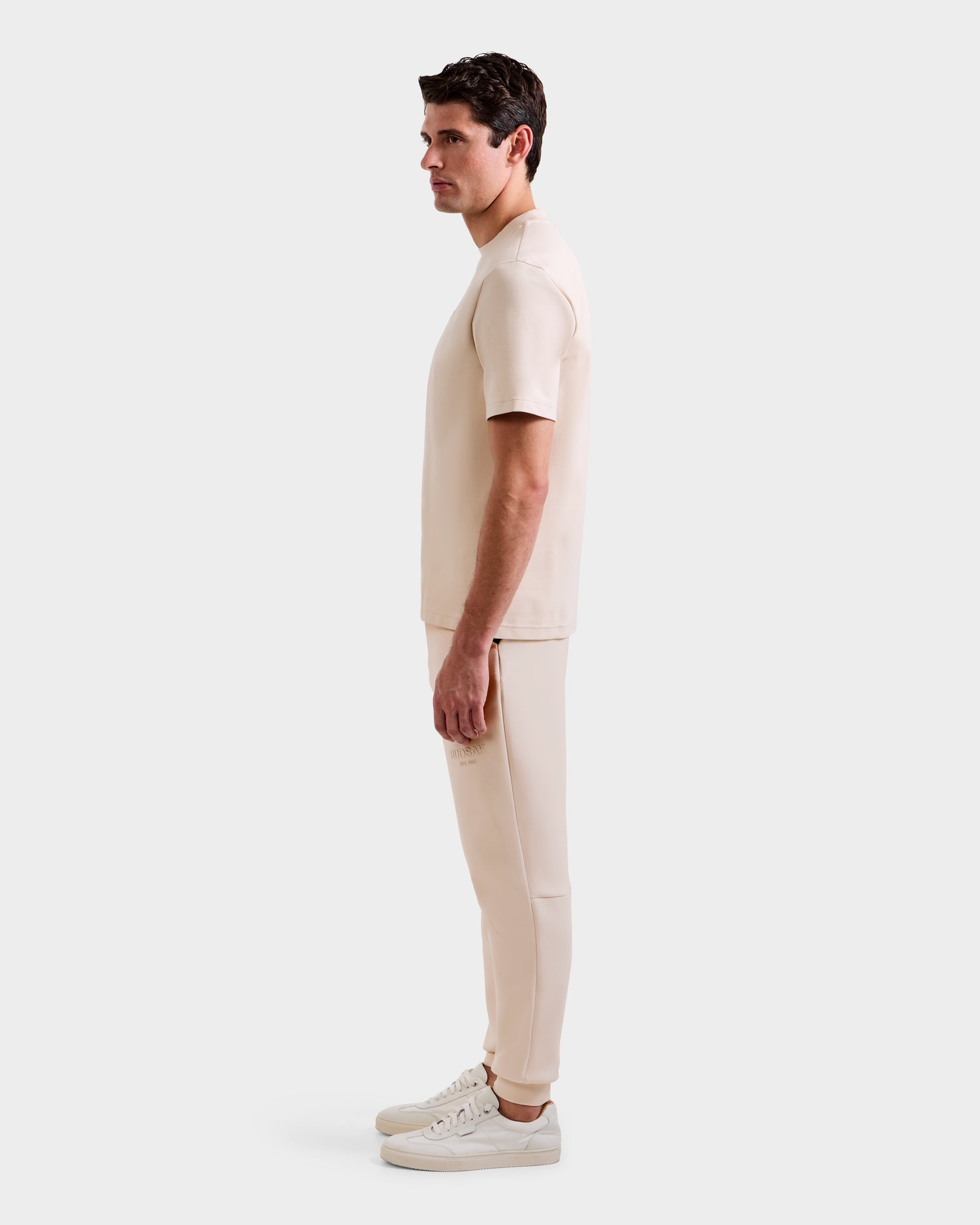 A man in profile, facing left, wears the Rudsak CASEY - 6525037 Vanilla men’s T-shirt and matching pants with white sneakers, set against a plain light gray background.