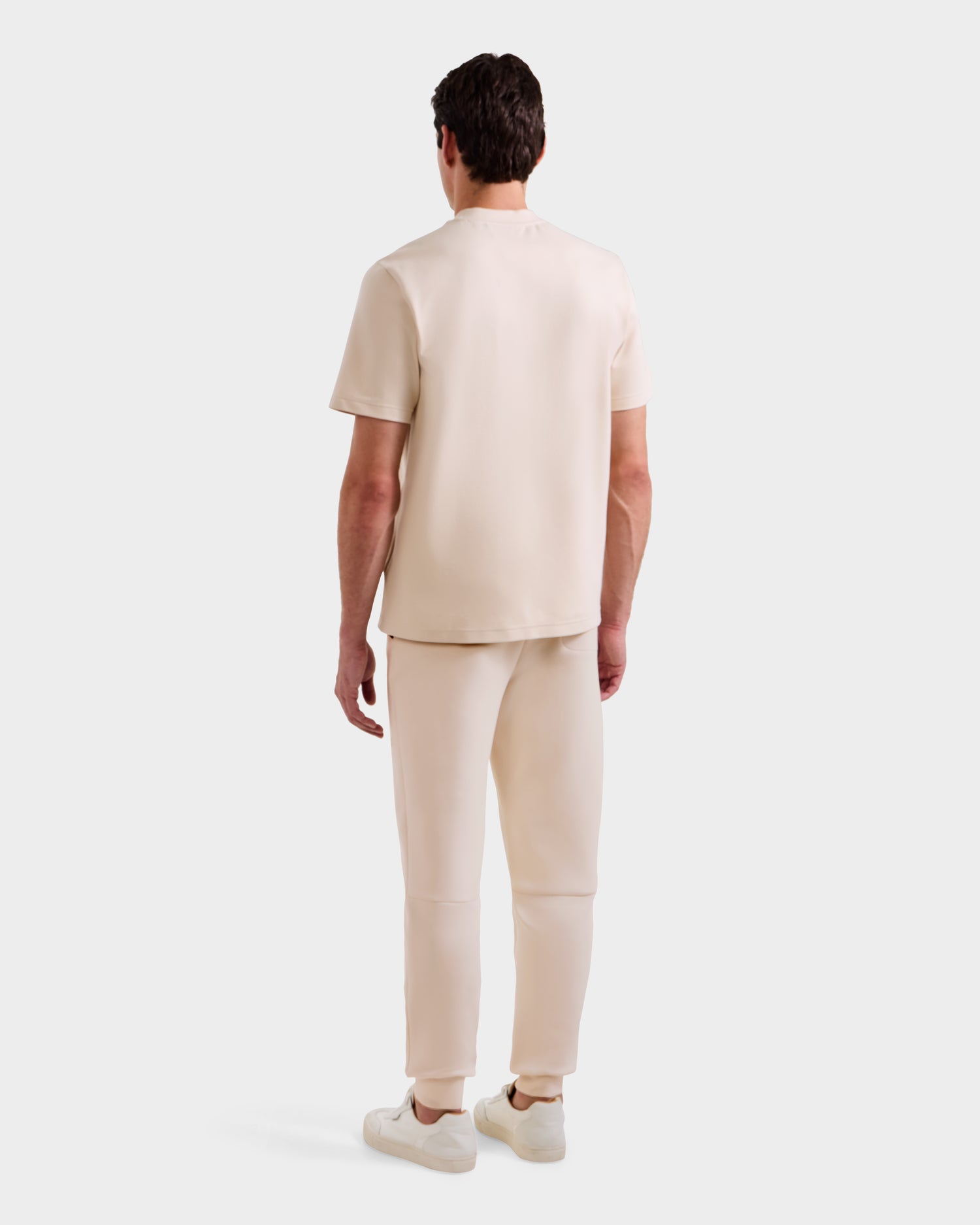 A person faces away, wearing the Rudsak CASEY - 6525037 Vanilla set—a comfortable, essential short-sleeve top and pants in light beige with white sneakers—against a plain light background.
