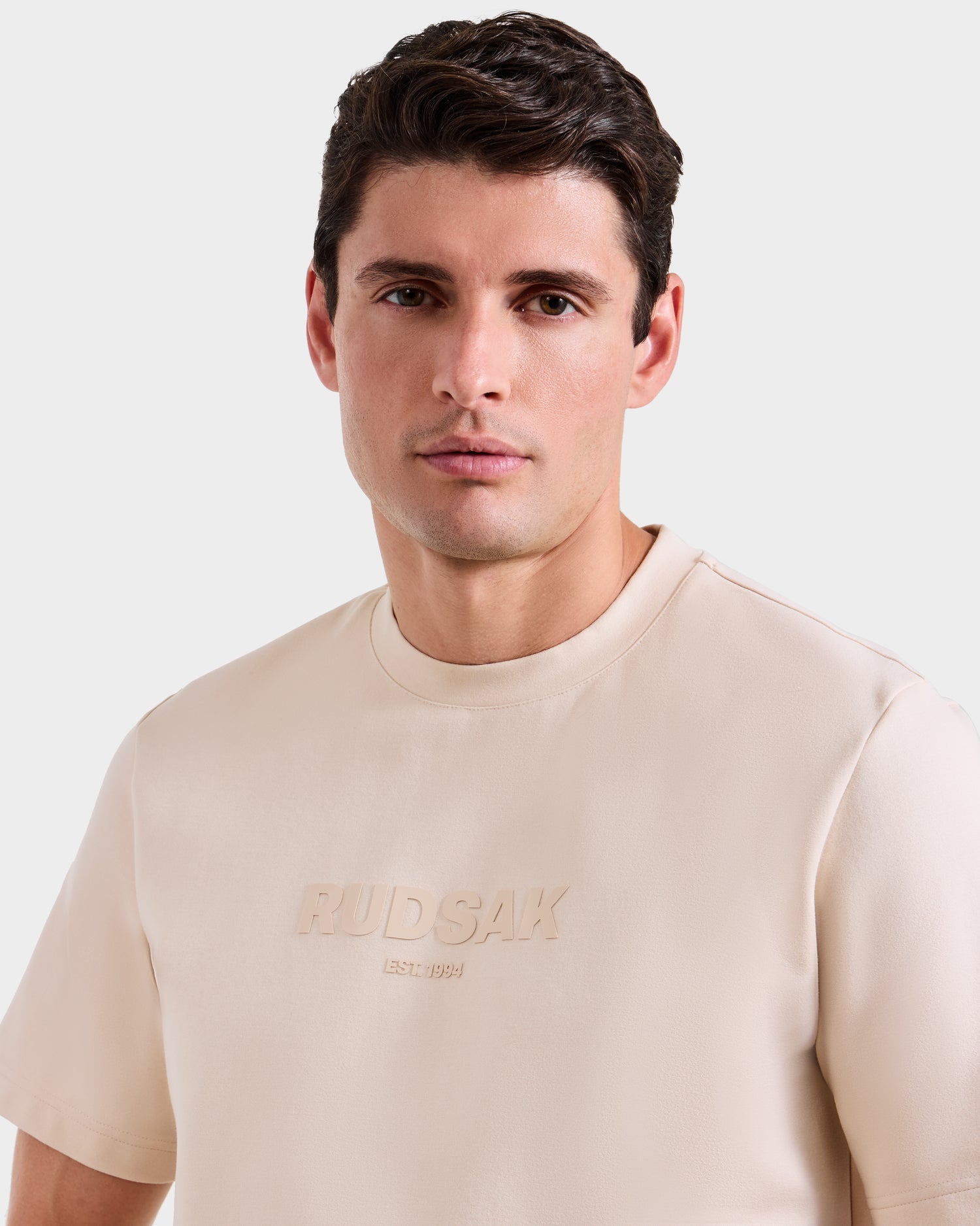 A man with short, dark hair wears the Rudsak CASEY - 6525037 Vanilla mens T-shirt with the brand name embossed on the front. He faces the camera neutrally against a plain light background.