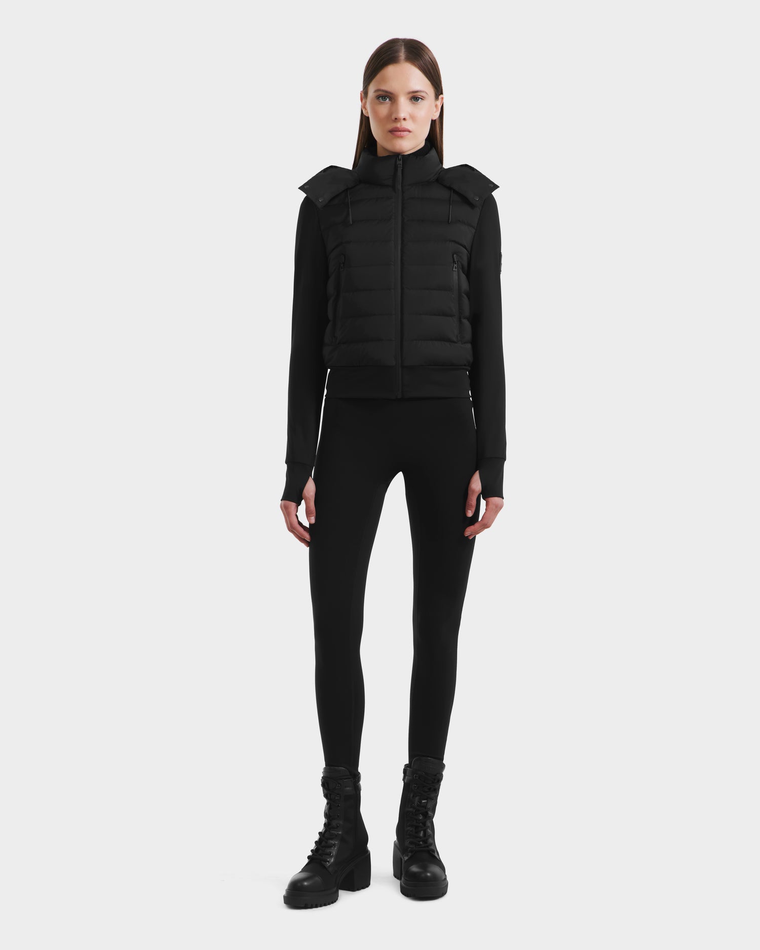 A woman faces forward in a Rudsak CASSA - 8125057 Black jacket, paired with black leggings and black lace-up boots, set against a plain white background.