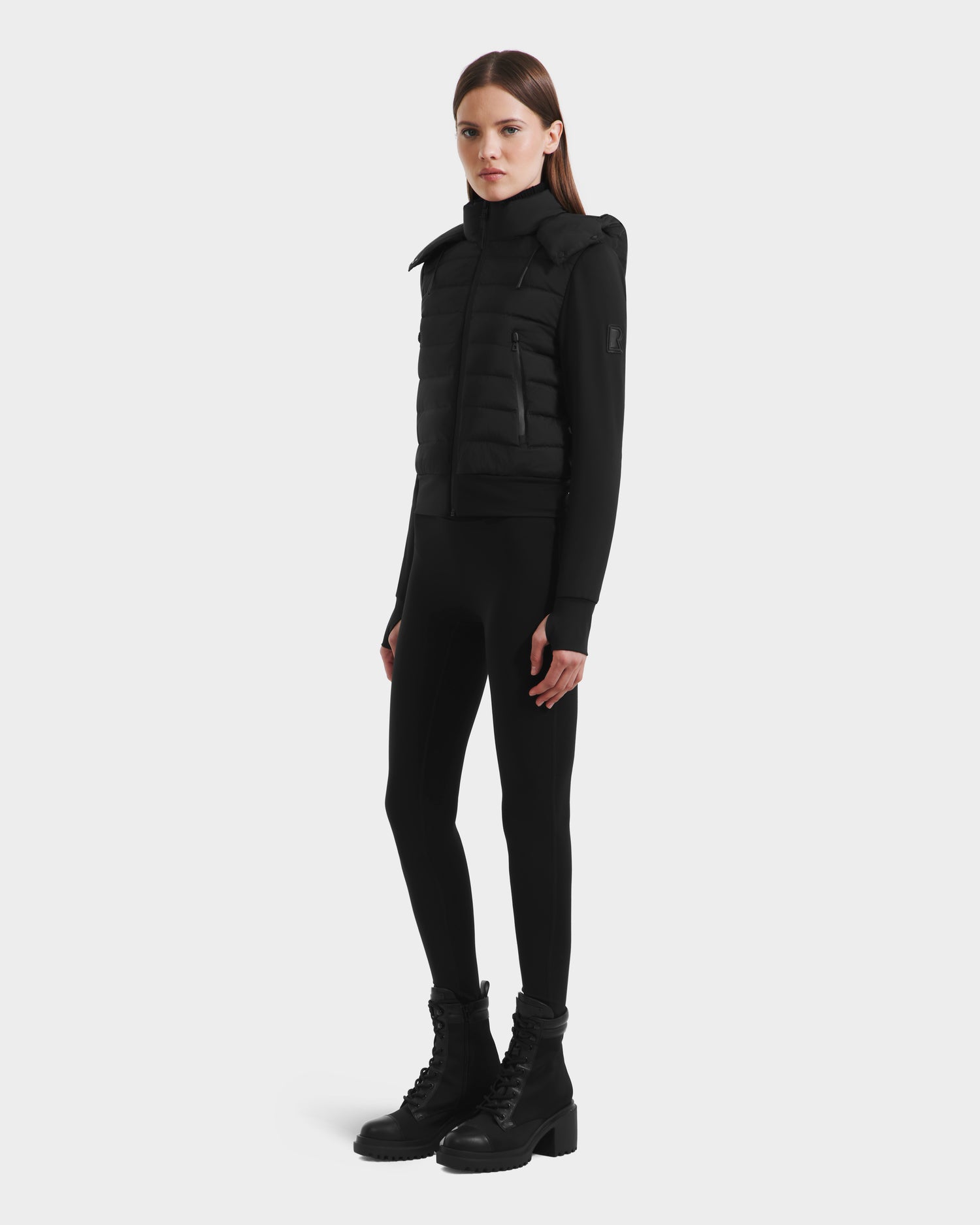 A woman with straight brown hair and a neutral expression stands against a white background, wearing the Rudsak CASSA - 8125057 Black transitional jacket over black leggings, a long-sleeve top, and lace-up boots.