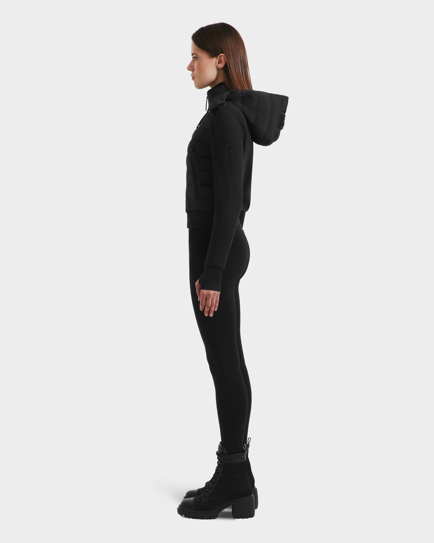 A woman in profile poses against a plain background, wearing the Rudsak CASSA - 8125057 Black transitional jacket with a hood, paired with black leggings and lace-up boots.
