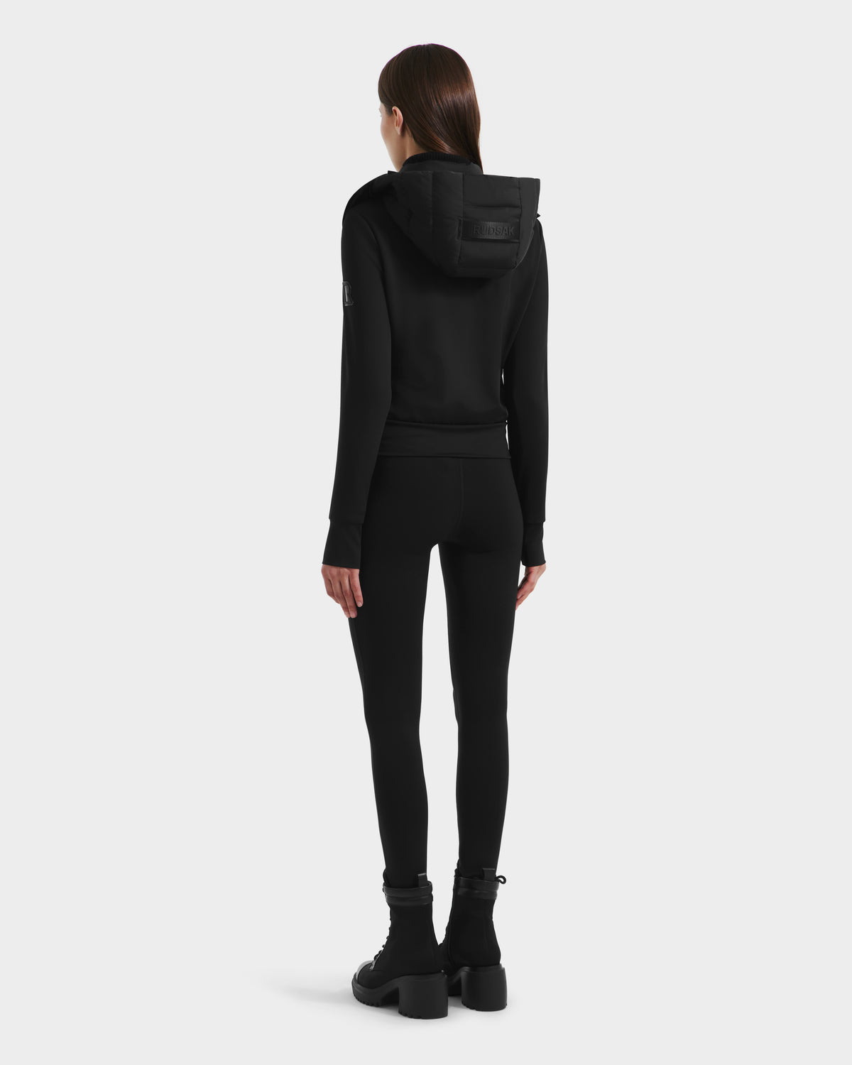A woman faces away, wearing the Rudsak CASSA - 8125057 Black women's transitional jacket with black leggings and boots against a plain white background.