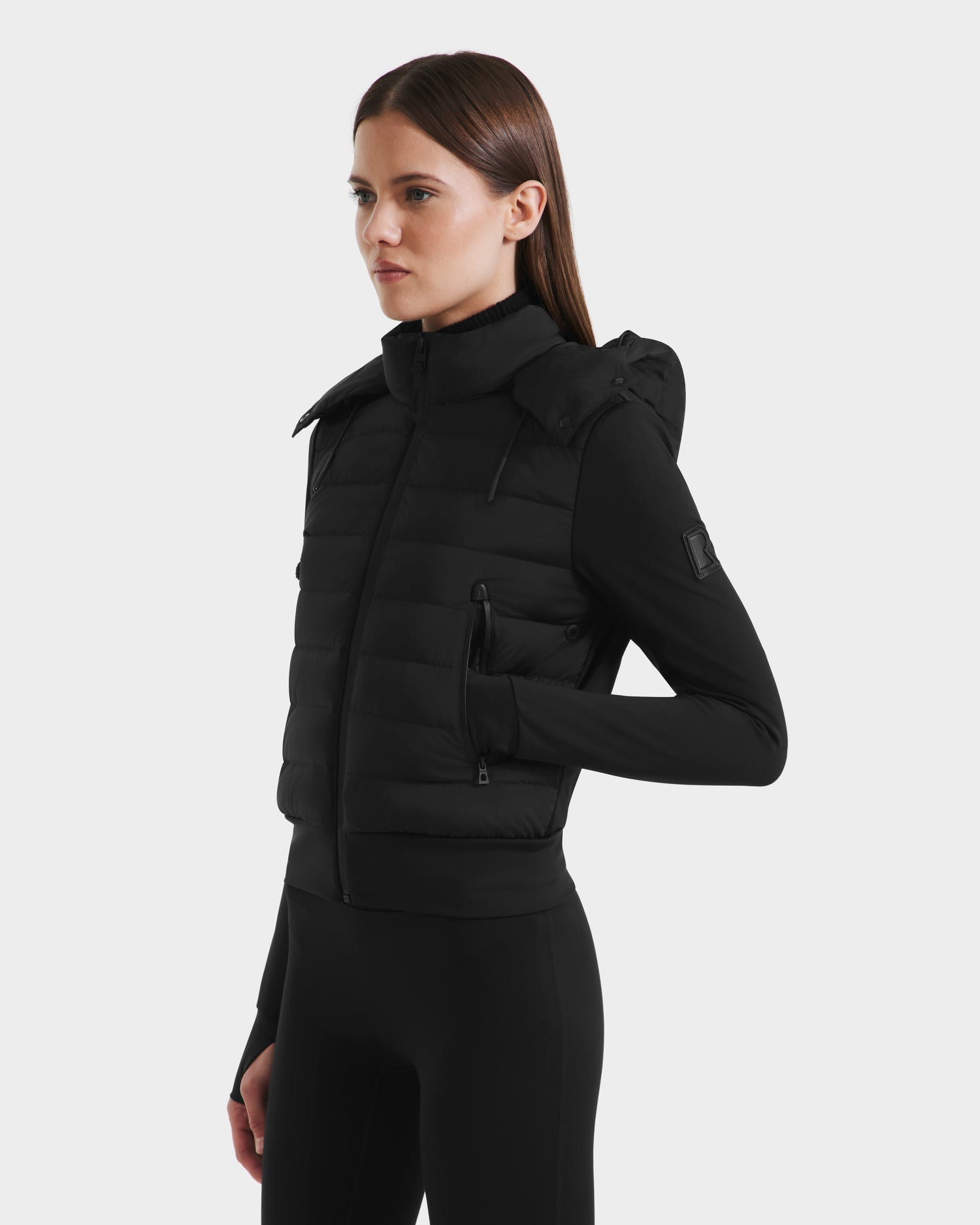 A woman with straight brown hair models the Rudsak CASSA - 8125057 Black transitional jacket, featuring a hood and front zipper, over a black long-sleeve top and pants, standing with one hand in her pocket against a light background.