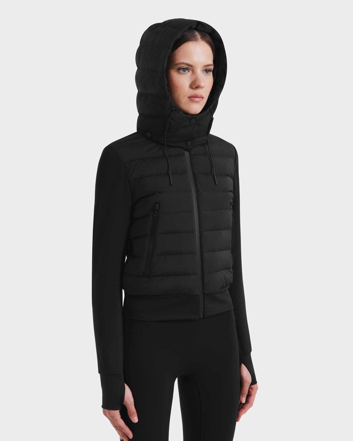 A woman wears the Rudsak CASSA - 8125057 Black fitted, lightweight cropped jacket with zippered pockets and matching black leggings, standing slightly to the side against a plain white background.