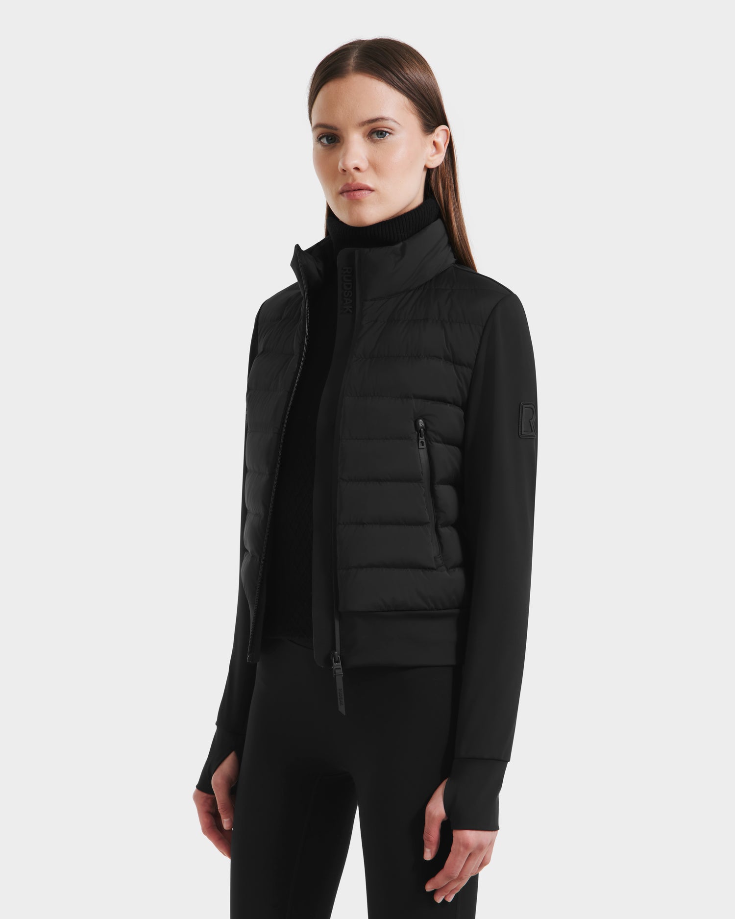A woman with straight brown hair wears the Rudsak CASSA - 8125057 Black lightweight cropped jacket over a black turtleneck and leggings, standing against a light gray background with a neutral expression—ideal for transitional weather.