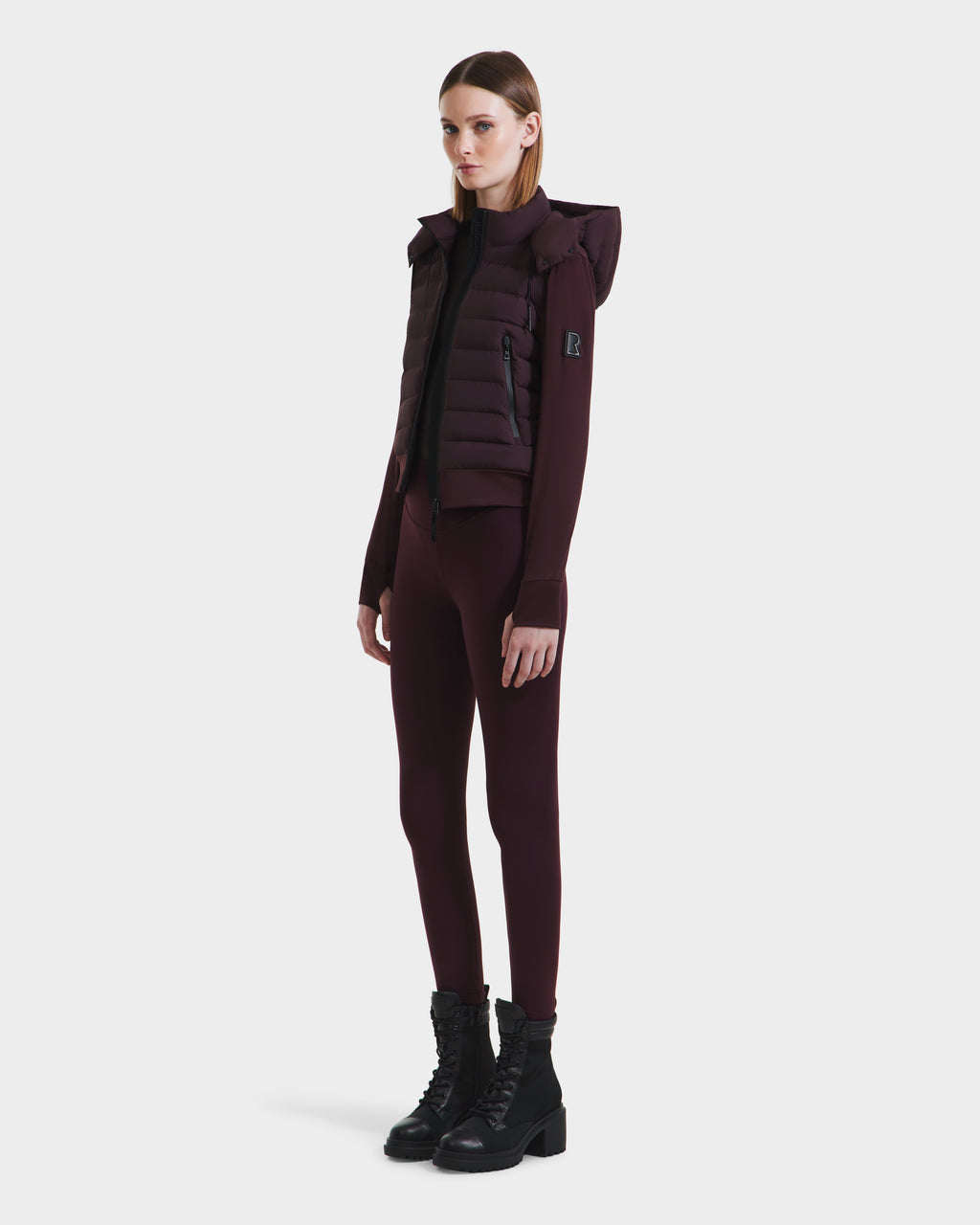 A woman wears the Rudsak CASSA - 8125057 Bordeaux puffer vest with a hood, styled with a matching fitted long-sleeve top, leggings, and black lace-up boots against a plain white background.