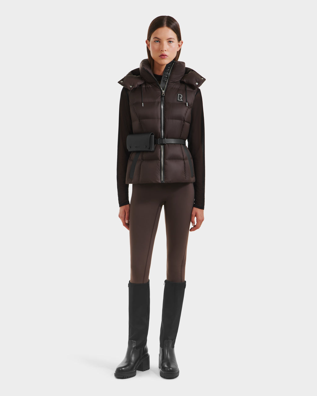 A woman stands facing forward against a white background, wearing the Rudsak CASSIDY N - 8125677 Espresso travel-friendly women’s down vest with a belt bag, styled with brown fitted pants, a black turtleneck, and knee-high black boots.
