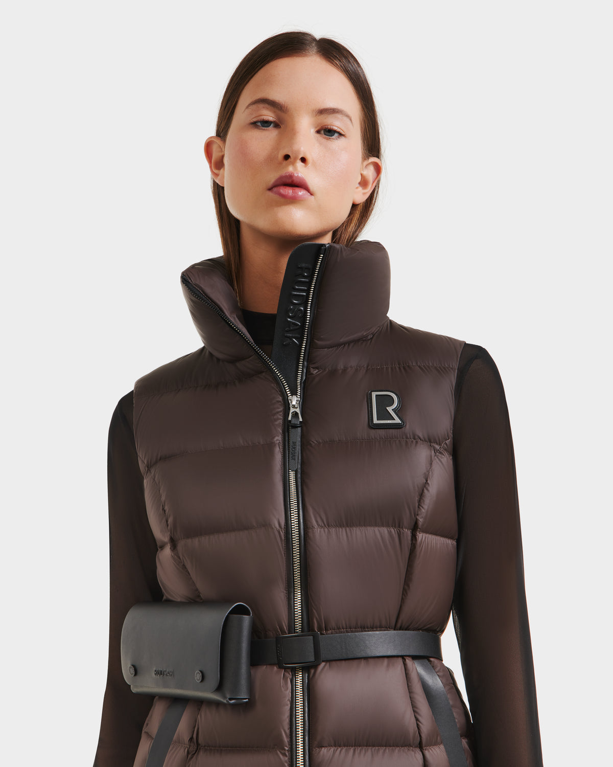 A woman wears the Rudsak CASSIDY N - 8125677 Espresso travel-friendly women’s down vest, featuring a high collar and lightweight warmth, styled with a black belt bag over a sheer black long-sleeve top against a plain white background.