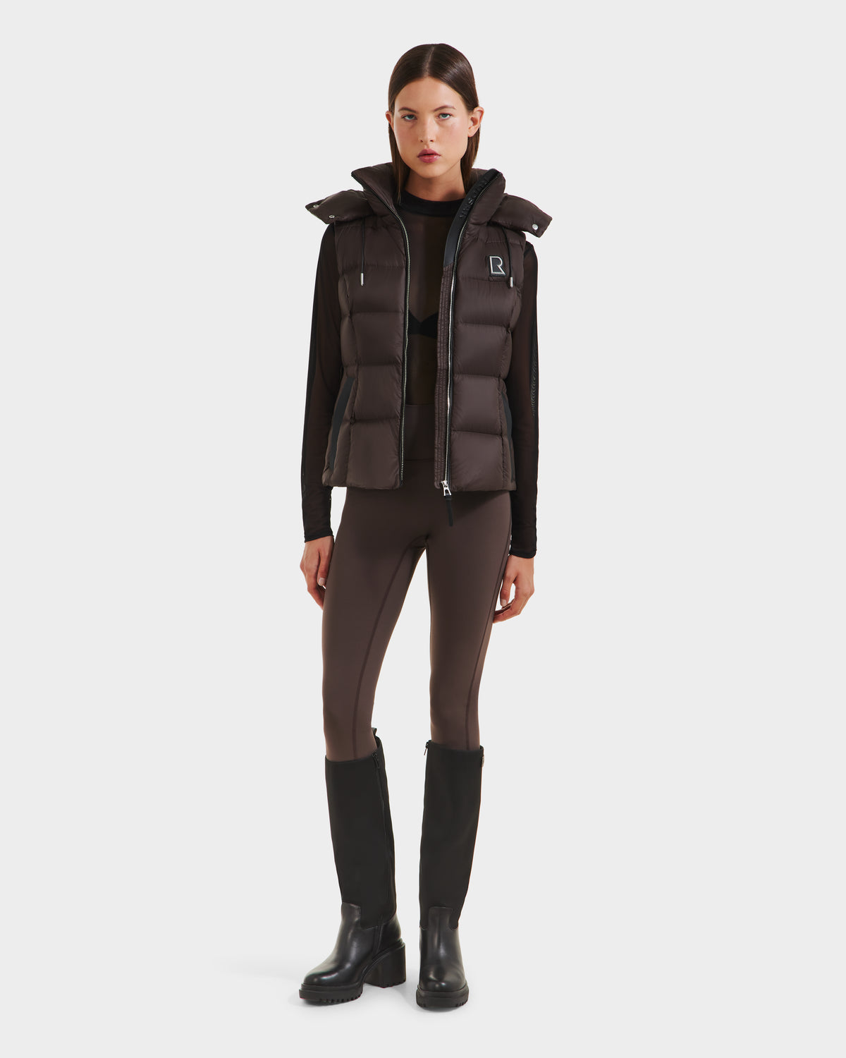 A woman faces forward, wearing the Rudsak CASSIDY N - 8125677 Espresso travel-friendly women’s down vest over a long-sleeve black top, brown leggings with black panels, and knee-high black boots against a plain white background.