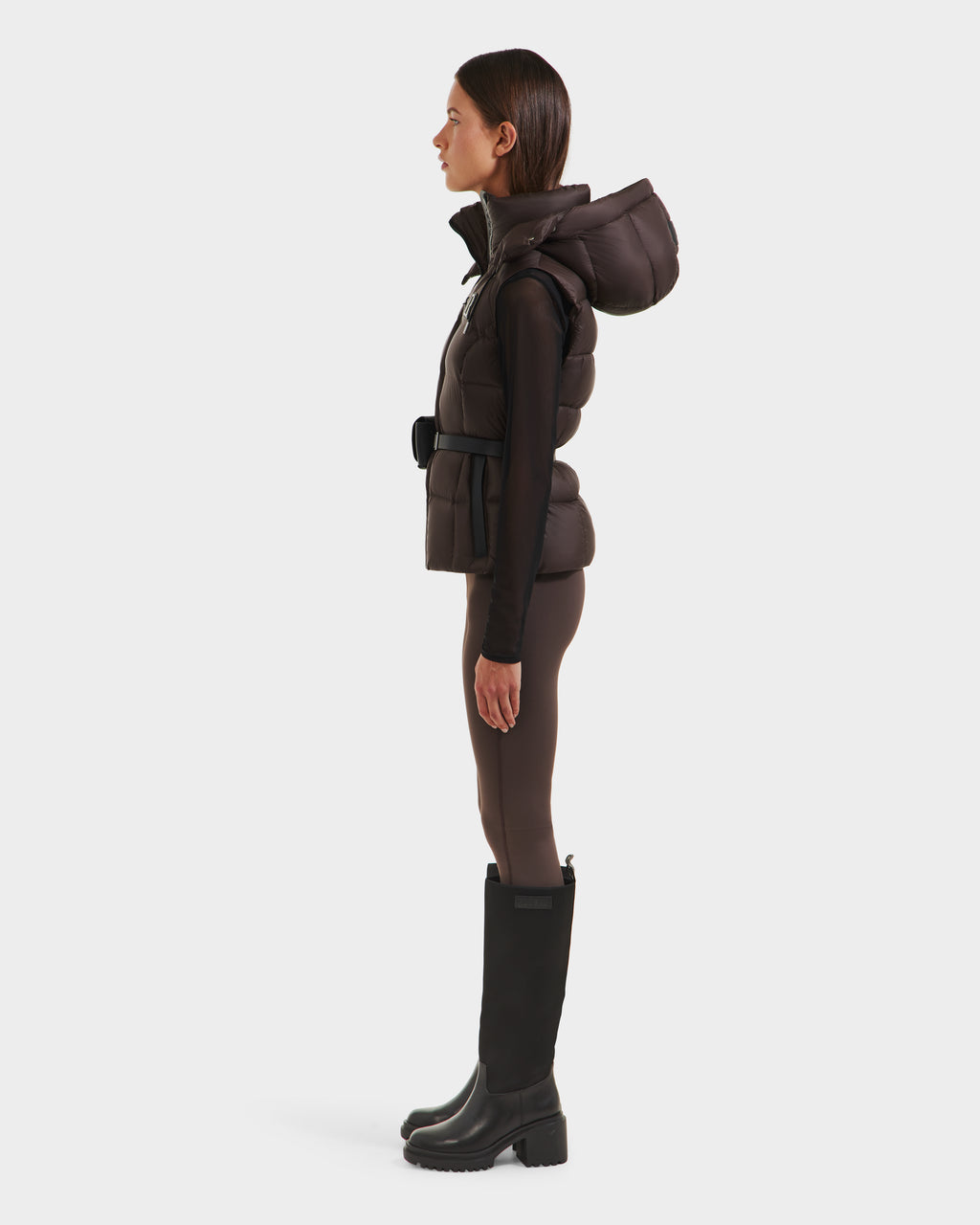 A woman stands in profile against a white background, wearing the Rudsak CASSIDY N - 8125677 Espresso down vest with hood and belted waist, paired with fitted pants and knee-high black boots.
