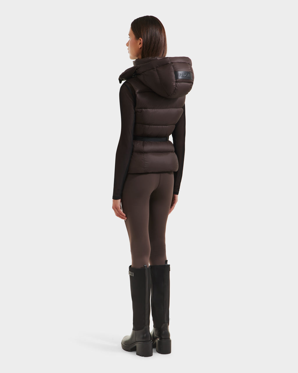 A woman faces away, wearing the Rudsak CASSIDY N - 8125677 Espresso women’s down vest over a black long-sleeve top, brown leggings, and black knee-high boots against a plain white background.