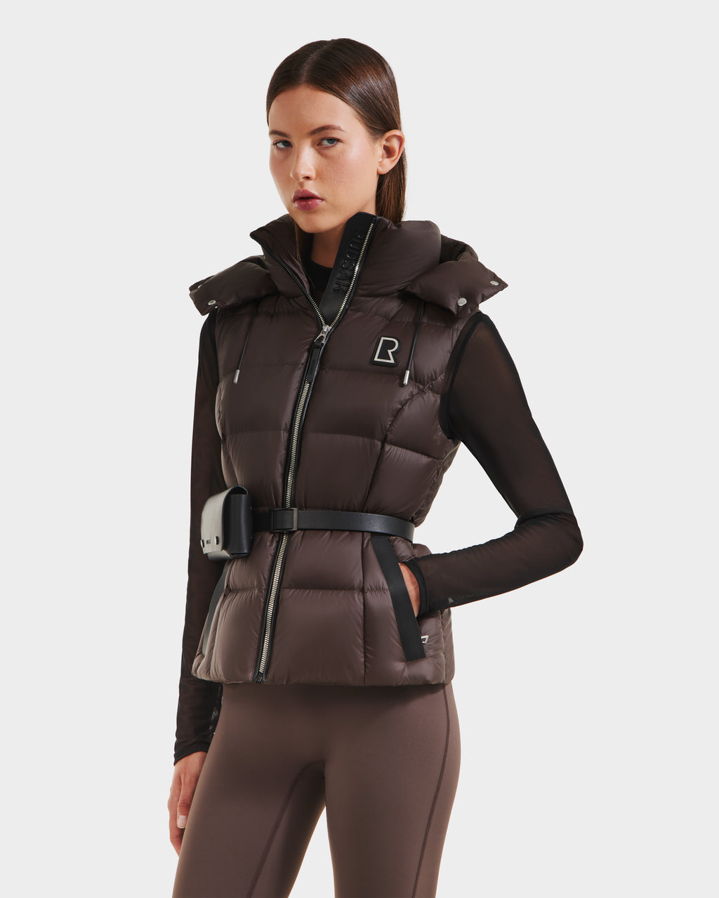 A woman models the Rudsak CASSIDY N - 8125677 Espresso fitted down vest with belted waist and pouch, made from recycled nylon shell, over a black long-sleeve top and brown leggings against a white background.