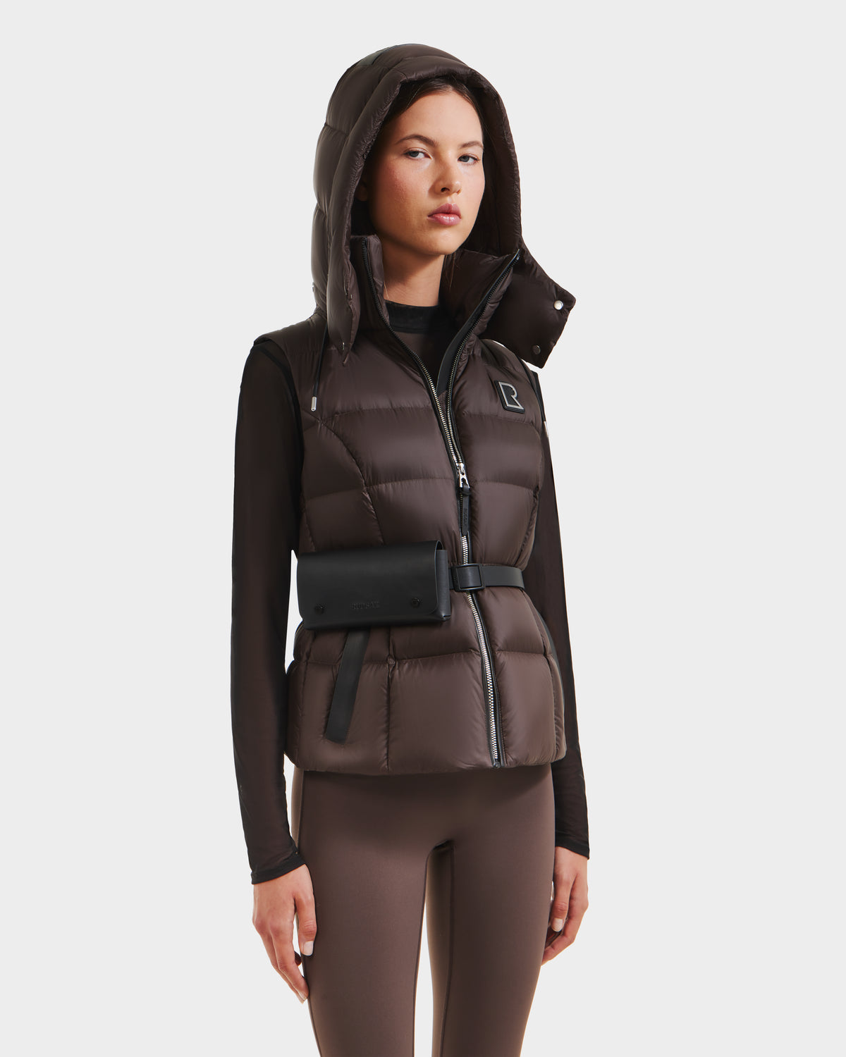 A woman wears the Rudsak CASSIDY N - 8125677 Espresso down vest with a hood, paired with a black belt bag, a fitted black long-sleeve top, and brown leggings, standing against a white background.