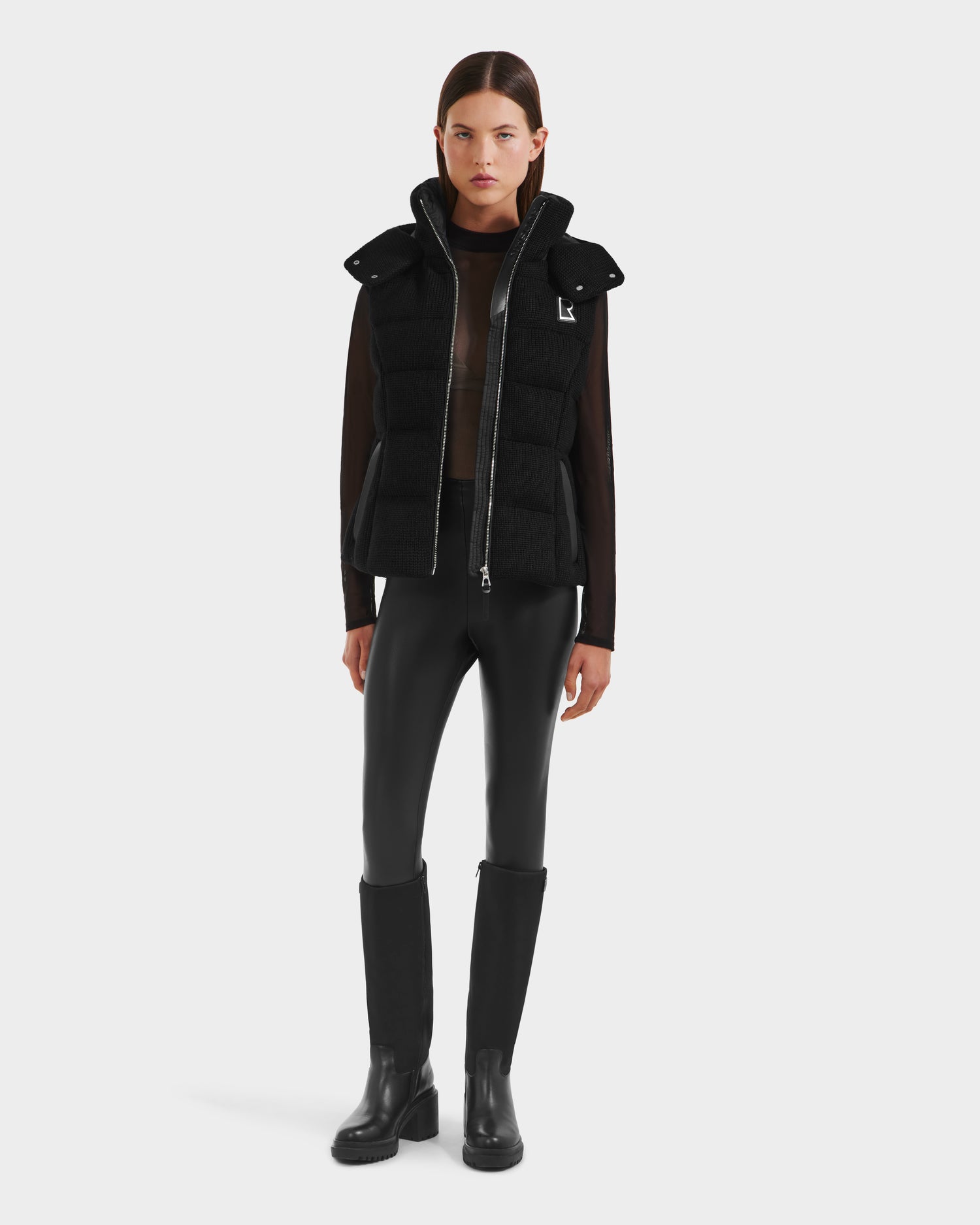 A woman faces forward against a plain white background, wearing the Rudsak CASSIDY - 8125622 Black women’s down vest, a sheer long-sleeve top, black fitted pants, and black knee-high boots.