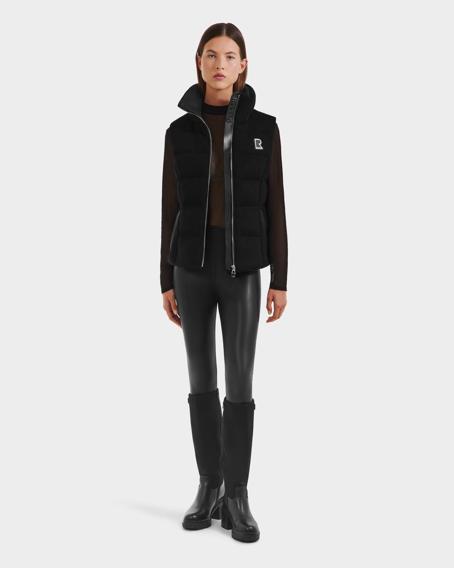 A woman stands facing forward in the Rudsak CASSIDY - 8125622 Black women’s down vest with removable hood, layered over a sheer black top, black leather pants, and knee-high boots against a plain white background.