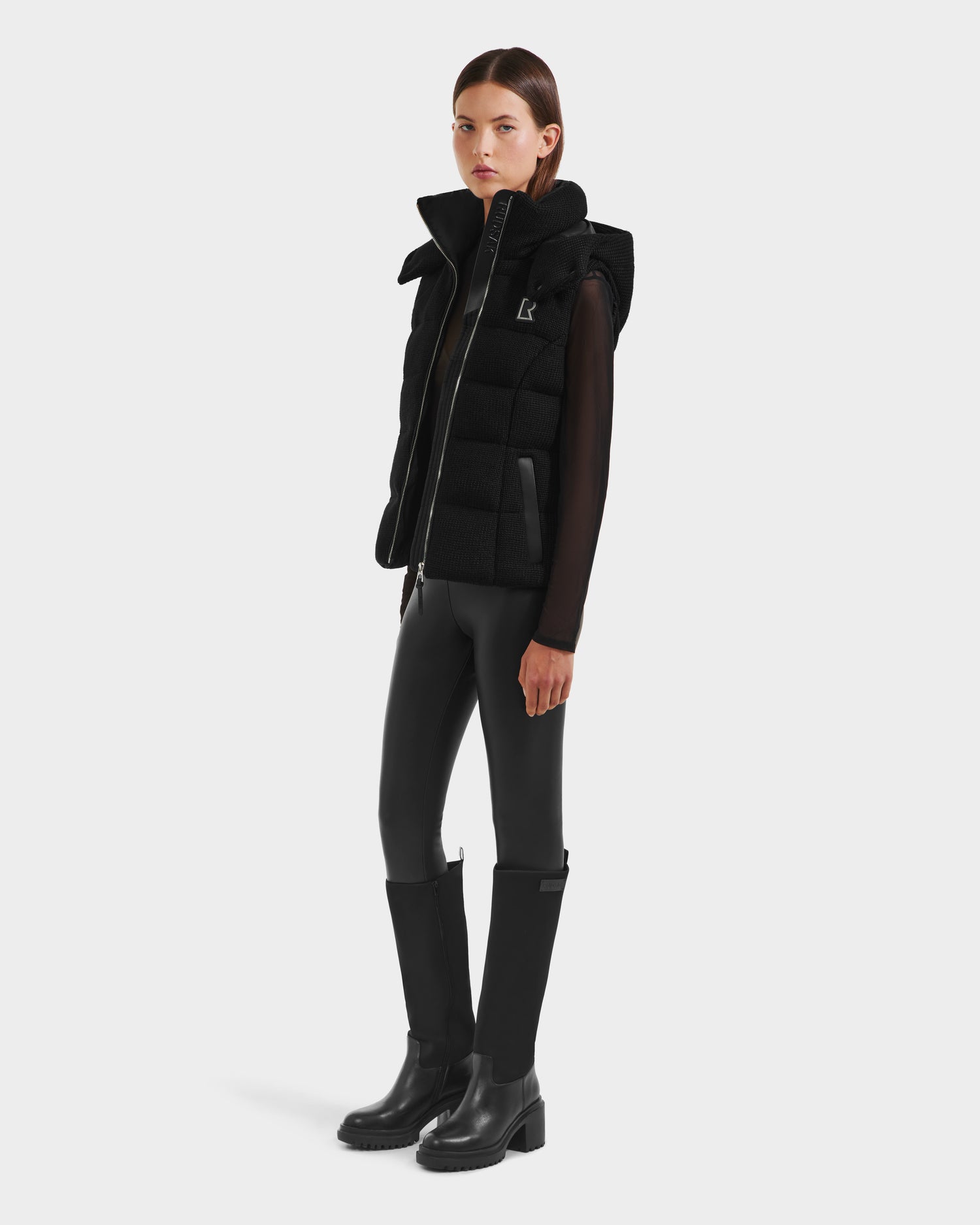 A woman stands facing forward in the Rudsak CASSIDY - 8125622 Black down vest, layered over a sheer long-sleeve top with black leather pants and knee-high boots, against a plain white background.