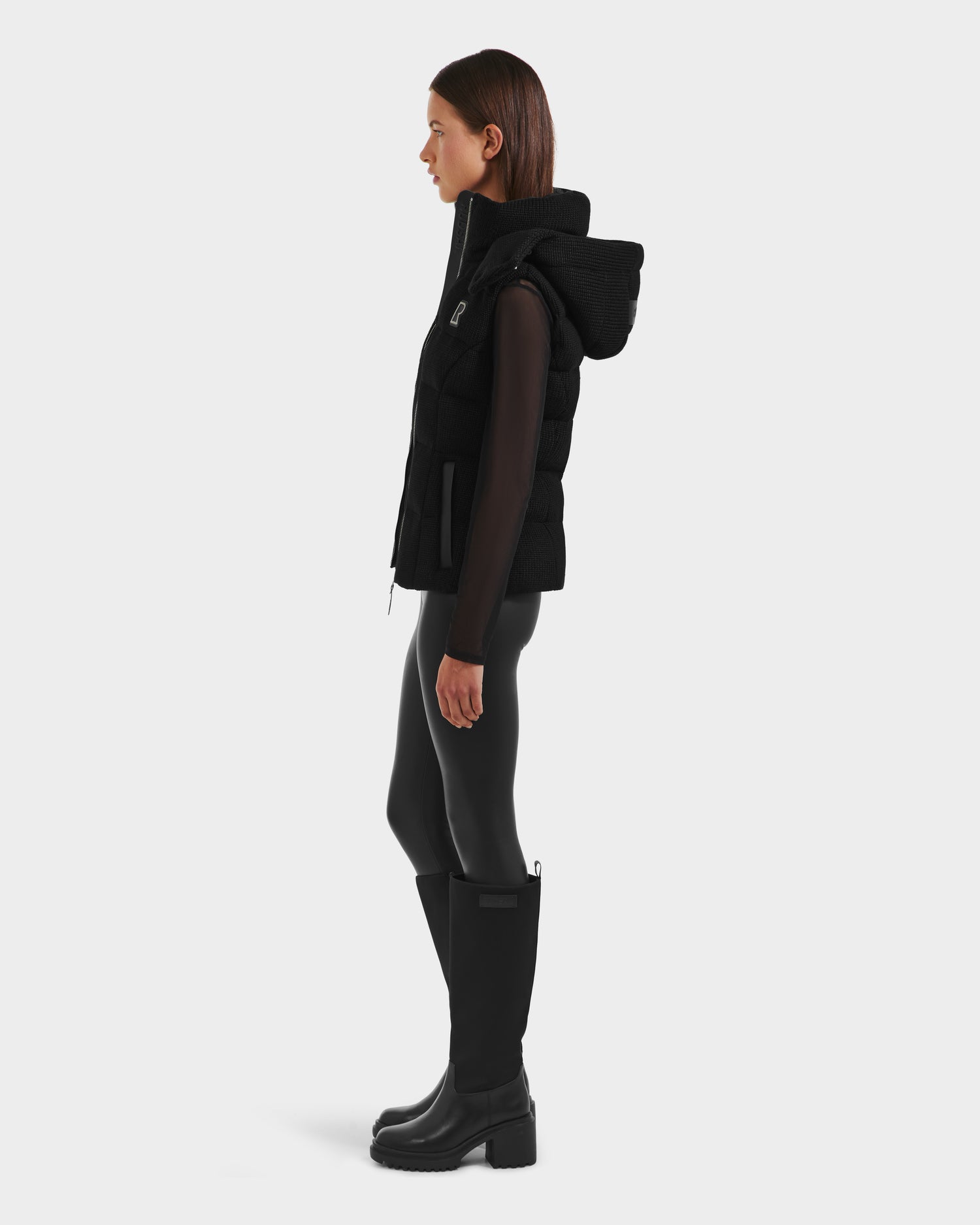 A woman stands in profile wearing the Rudsak CASSIDY - 8125622 Black womens down vest with a removable hood, sheer long-sleeve top, shiny leggings, and knee-high black boots, blending lightweight warmth with sleek style.