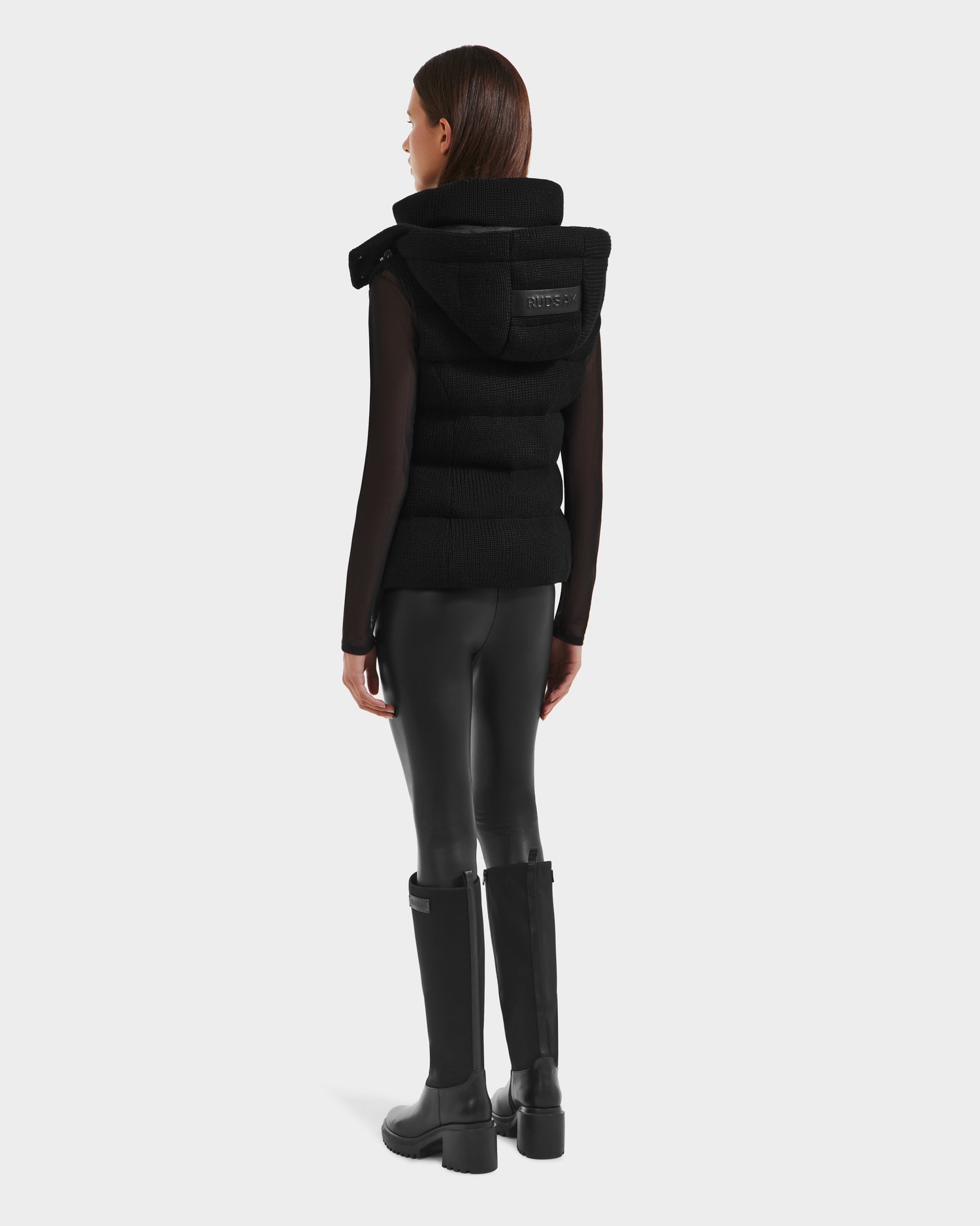 A woman faces away, wearing the Rudsak CASSIDY - 8125622 Black women’s down vest with removable hood, a black long-sleeve top, black faux leather leggings, and knee-high black boots against a light gray background.
