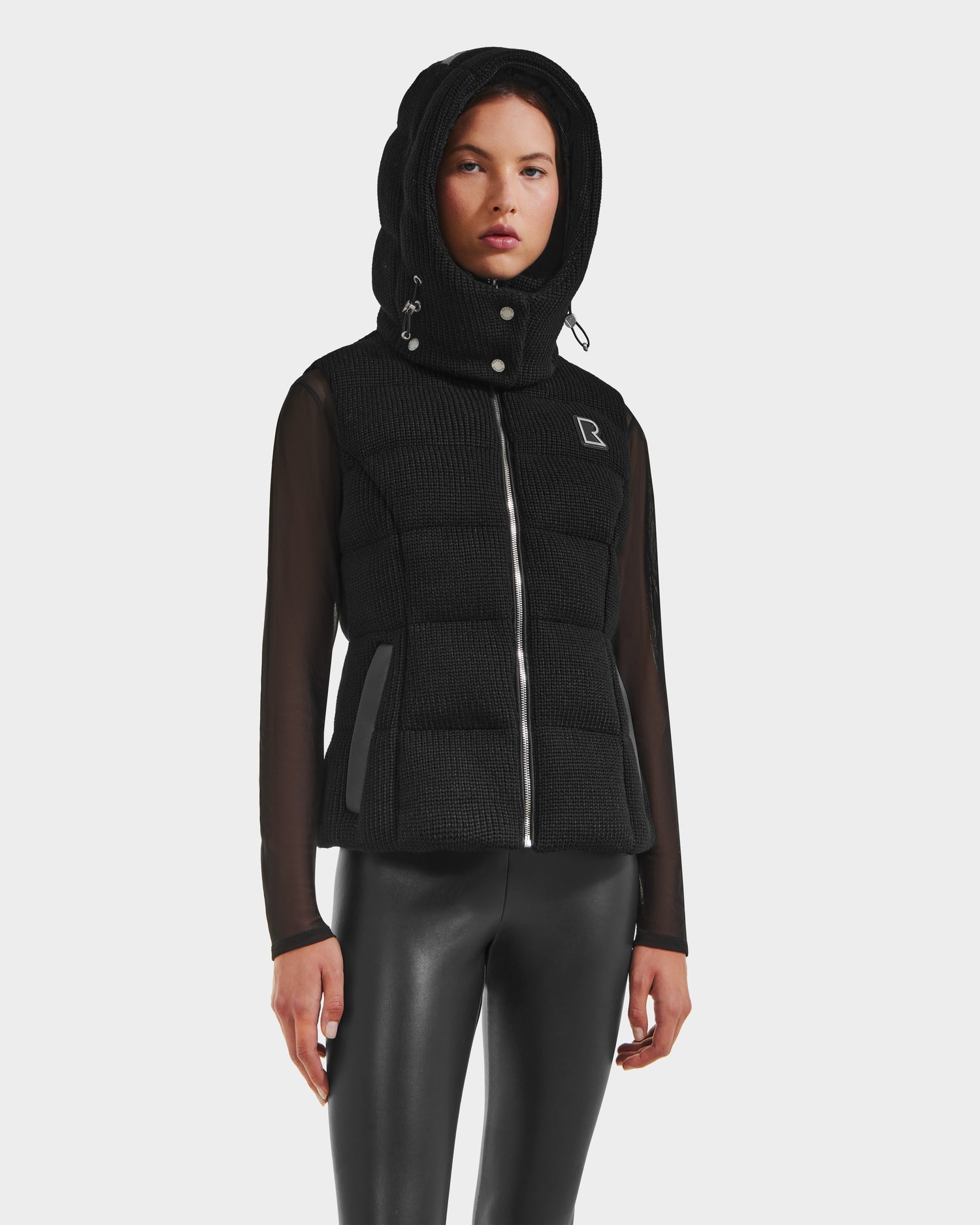 A woman models the Rudsak CASSIDY - 8125622 Black womens down vest with mesh long sleeves and a front zipper, paired with black leather pants against a white background, highlighting its lightweight warmth and versatile style.