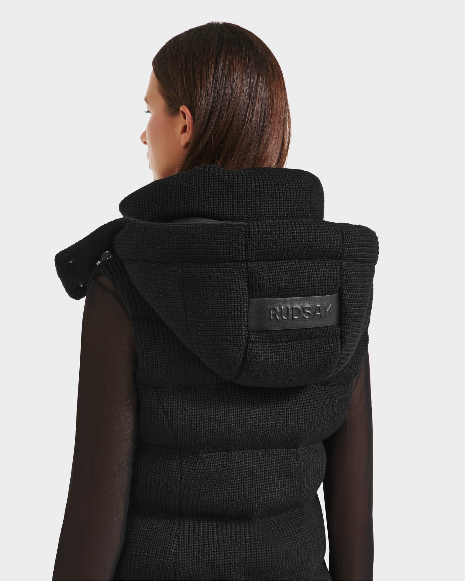 A woman with straight brown hair is shown from behind against a white background, wearing the Rudsak CASSIDY - 8125622 Black womens down vest with a removable hood and a visible Rudsak label on the back.
