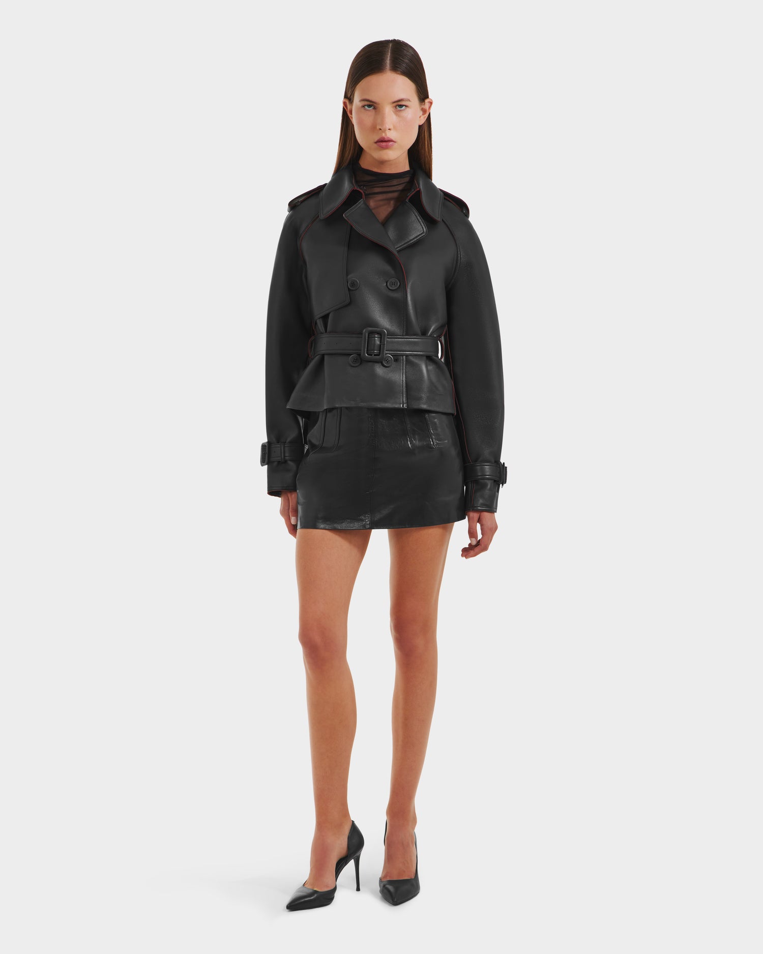 A woman faces forward in the CASSIE S-B - 8125554 Black double-breasted belted leather jacket by Rudsak, paired with a matching mini skirt, sheer black top, and high heels against a plain white background.