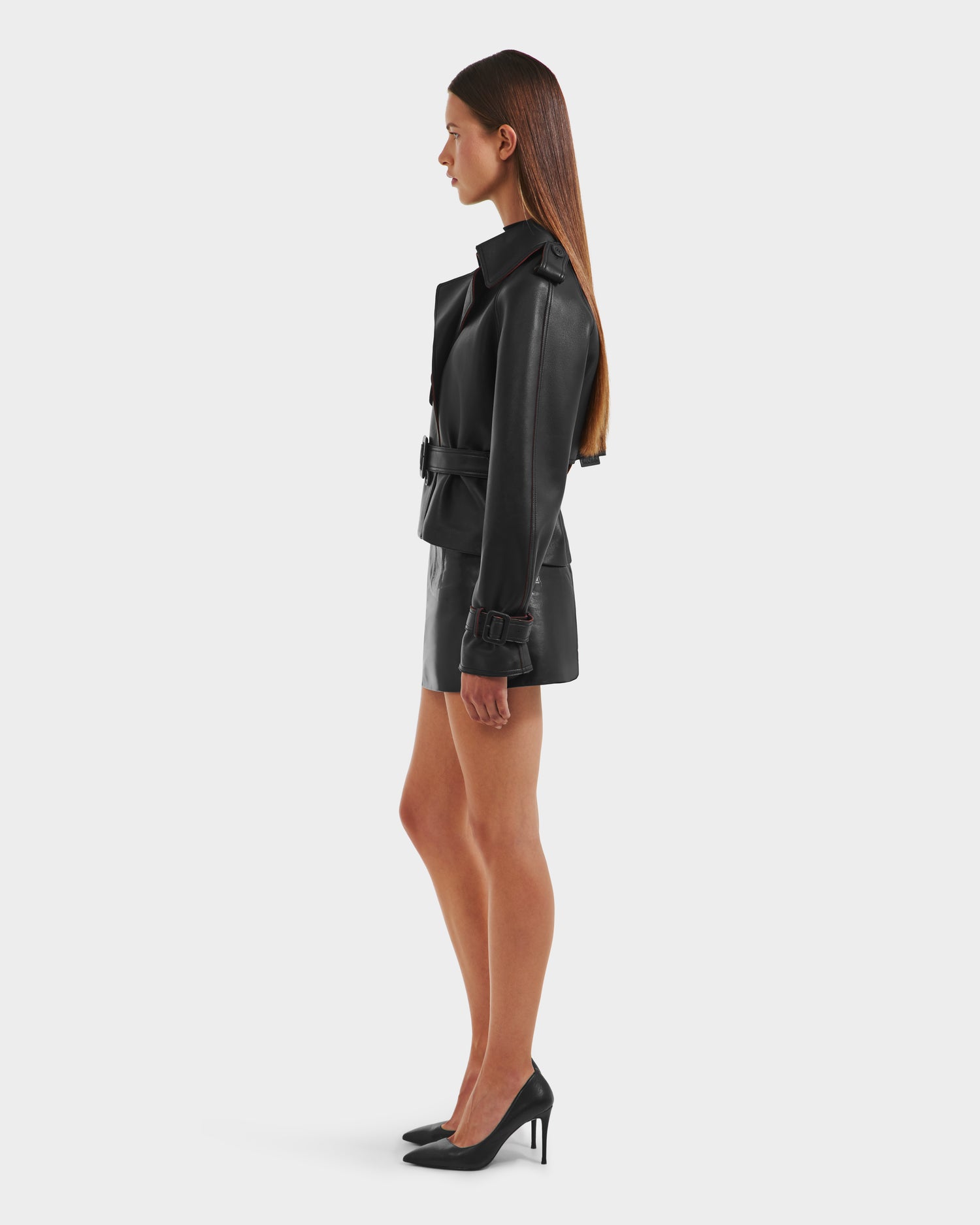 A woman in profile wears the Rudsak CASSIE S-B - 8125554 Black double-breasted faux leather jacket and a black mini skirt with high heels against a plain white background.