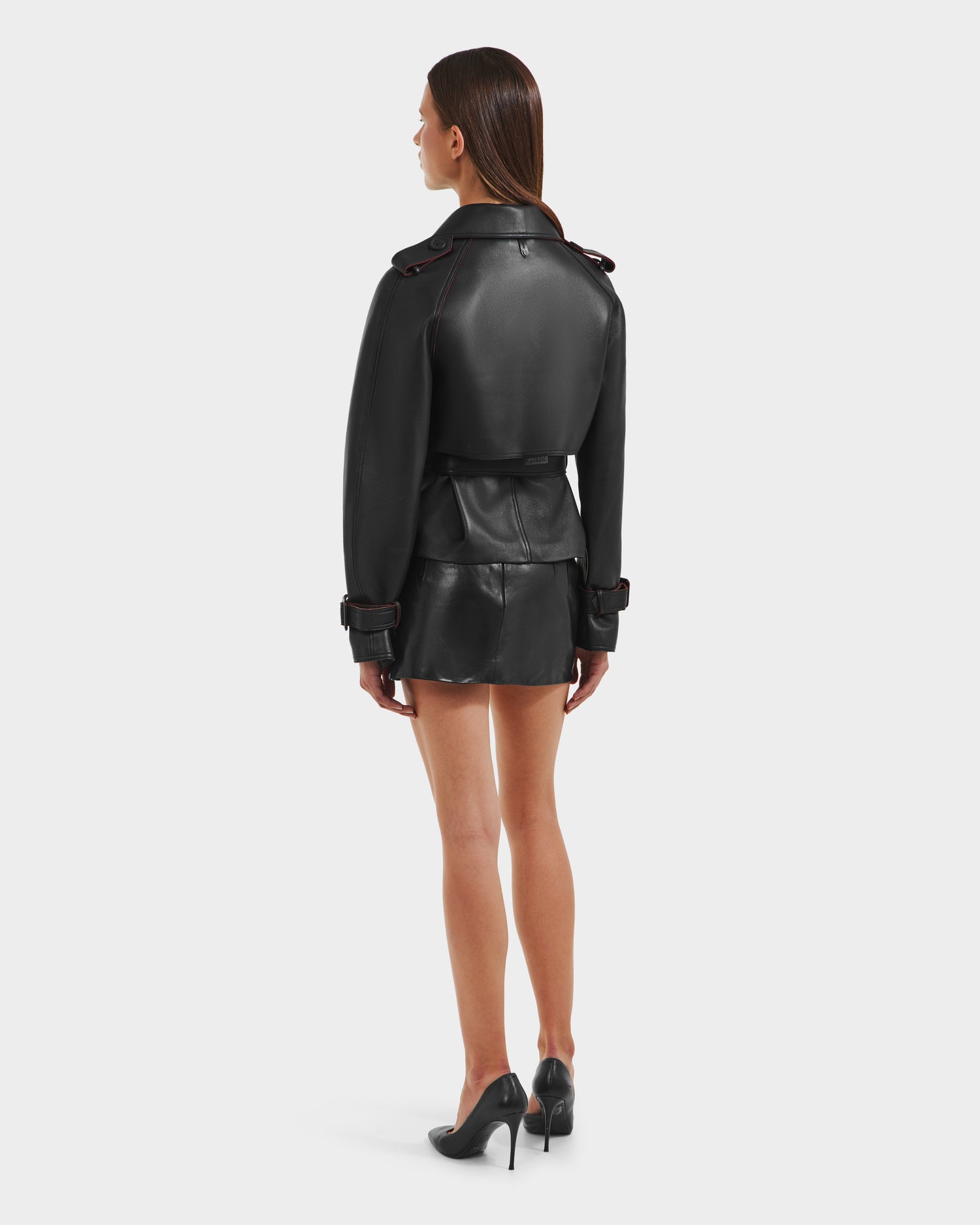 A woman faces away in the Rudsak CASSIE S-B - 8125554 Black double-breasted leather jacket and matching shorts, paired with black heels. Her straight brown hair is down as she stands against a plain white background.