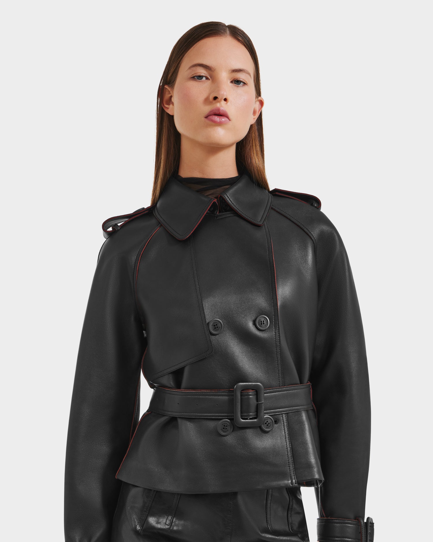 A woman with straight brown hair wears the Rudsak CASSIE S-B - 8125554 black double-breasted bonded leather jacket with a belt, button details, and wide collar, standing against a plain light background.