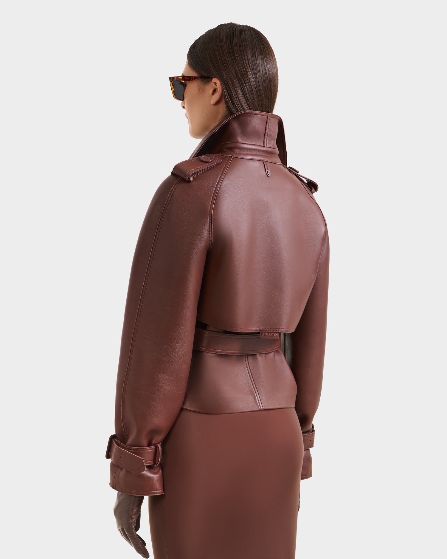 A woman shown from the back stands against a white background wearing Rudsak’s CASSIE S-B - 8125554 Rust: a cropped bonded leather trench with high collar and belt, paired with matching brown skirt, gloves, and RUDSAK sunglasses.
