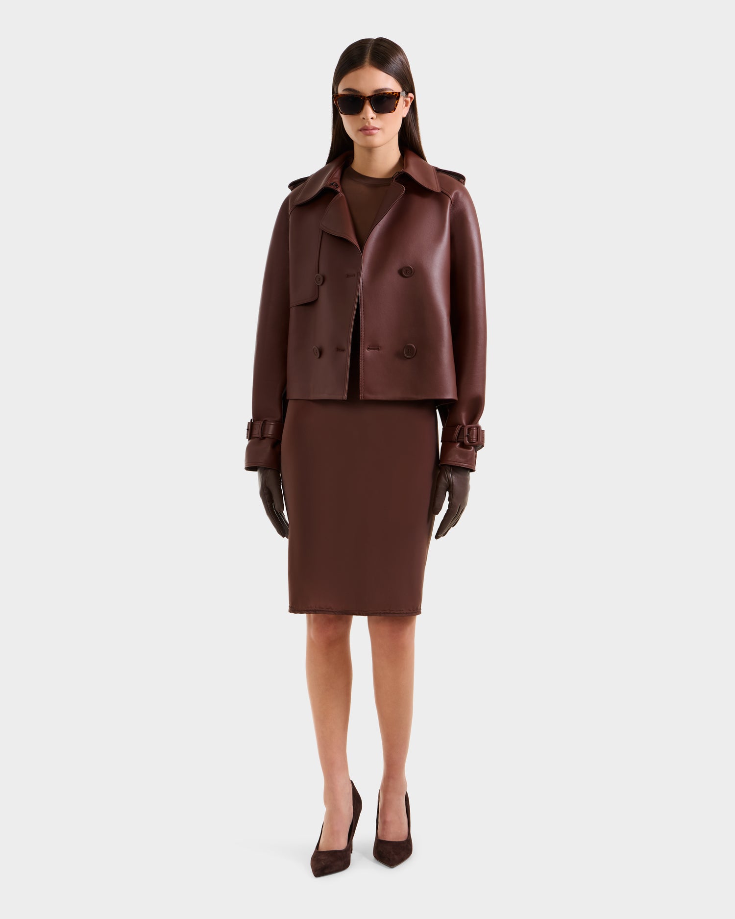 A woman faces forward against a light background, wearing the Rudsak CASSIE S-B - 8125554 Rust bonded leather jacket, a matching knee-length dress, dark gloves, pointed heels, and oversized sunglasses.