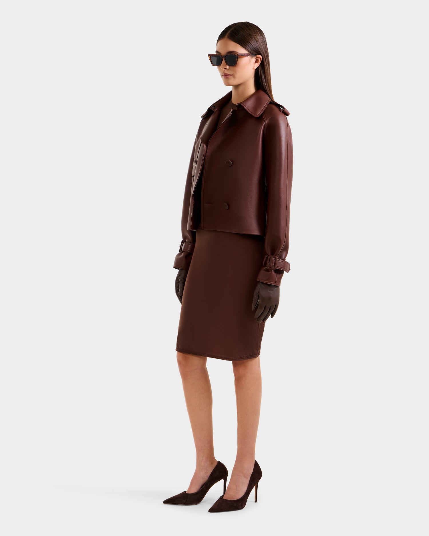 A woman in dark sunglasses wears the Rudsak CASSIE S-B - 8125554 Rust bonded leather jacket with a matching skirt, gloves, and high heels. She stands slightly turned to the right against a plain light gray background.