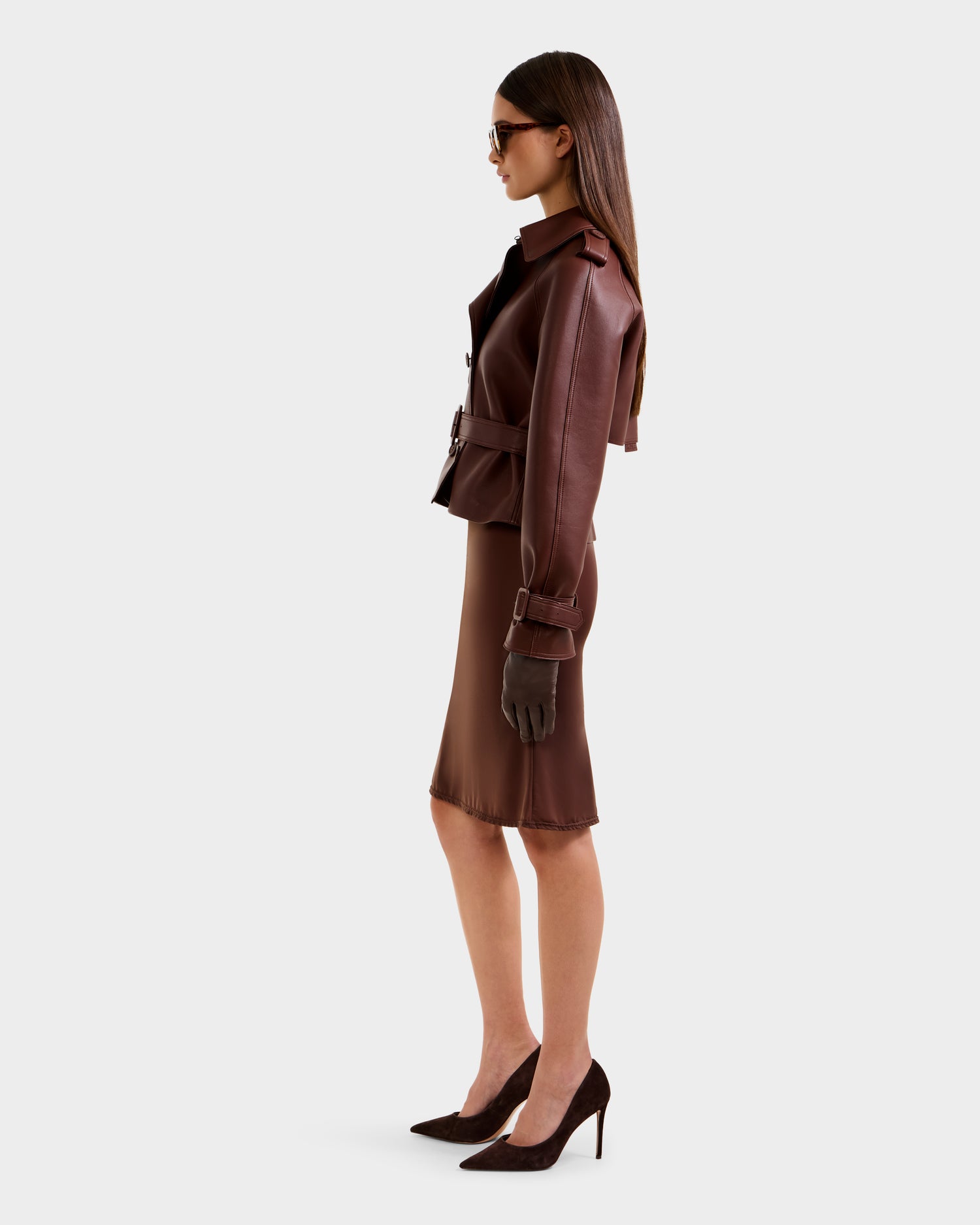 A woman stands in profile against a white background, wearing the Rudsak CASSIE S-B - 8125554 Rust jacket with a brown skirt, matching heels, gloves, and sunglasses.