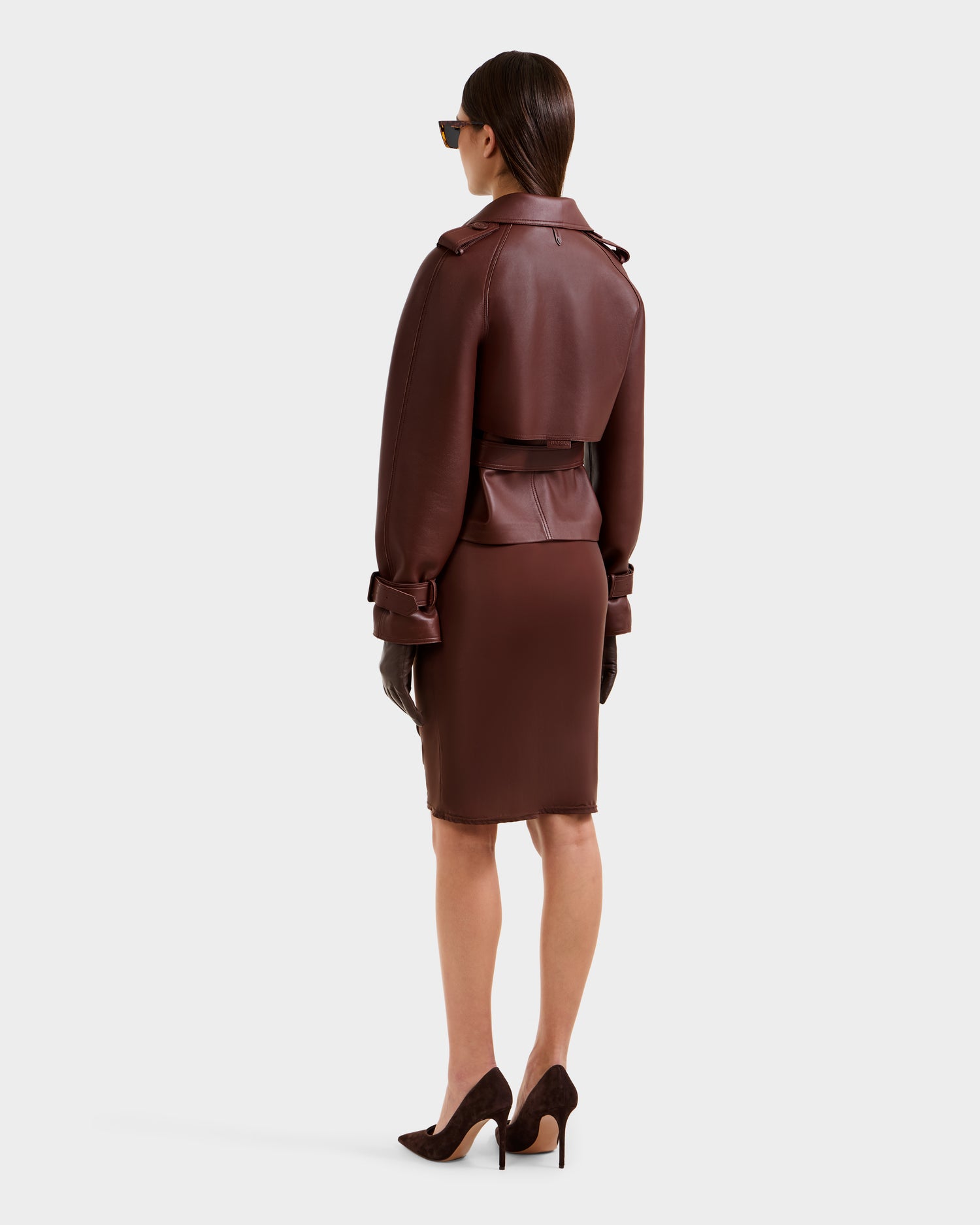 A woman faces away, wearing the Rudsak CASSIE S-B (8125554 Rust) cropped trench coat in brown bonded leather, paired with a matching knee-length skirt, brown heels, dark gloves, sunglasses, and straight dark hair tucked behind her ears.