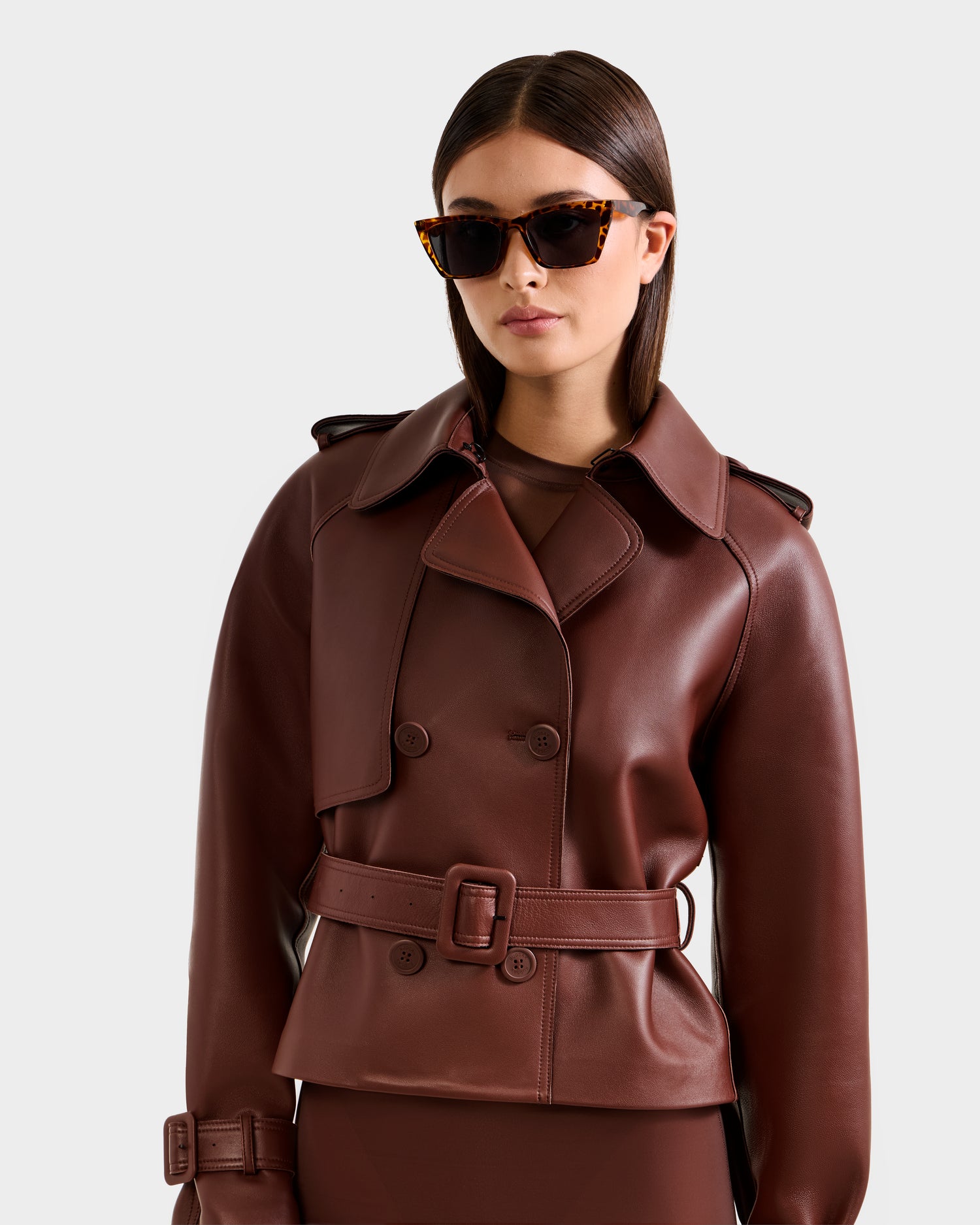 A woman in brown tortoiseshell sunglasses and the Rudsak CASSIE S-B - 8125554 Rust belted, double-breasted trench coat stands against a plain light background, gazing confidently at the camera.