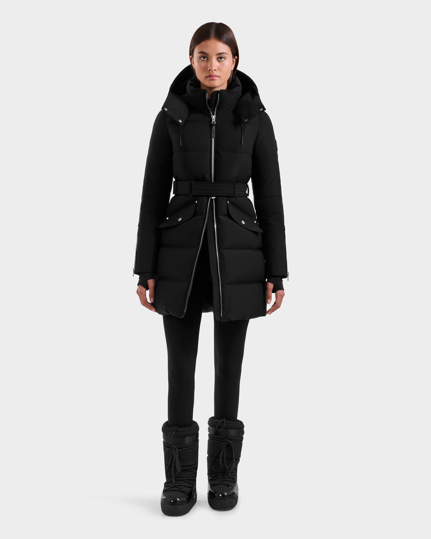 A person stands facing forward, wearing the Rudsak CAT H - 8125588 Black women’s down puffer coat with a belt, black leggings, and chunky black winter boots against a plain light background.