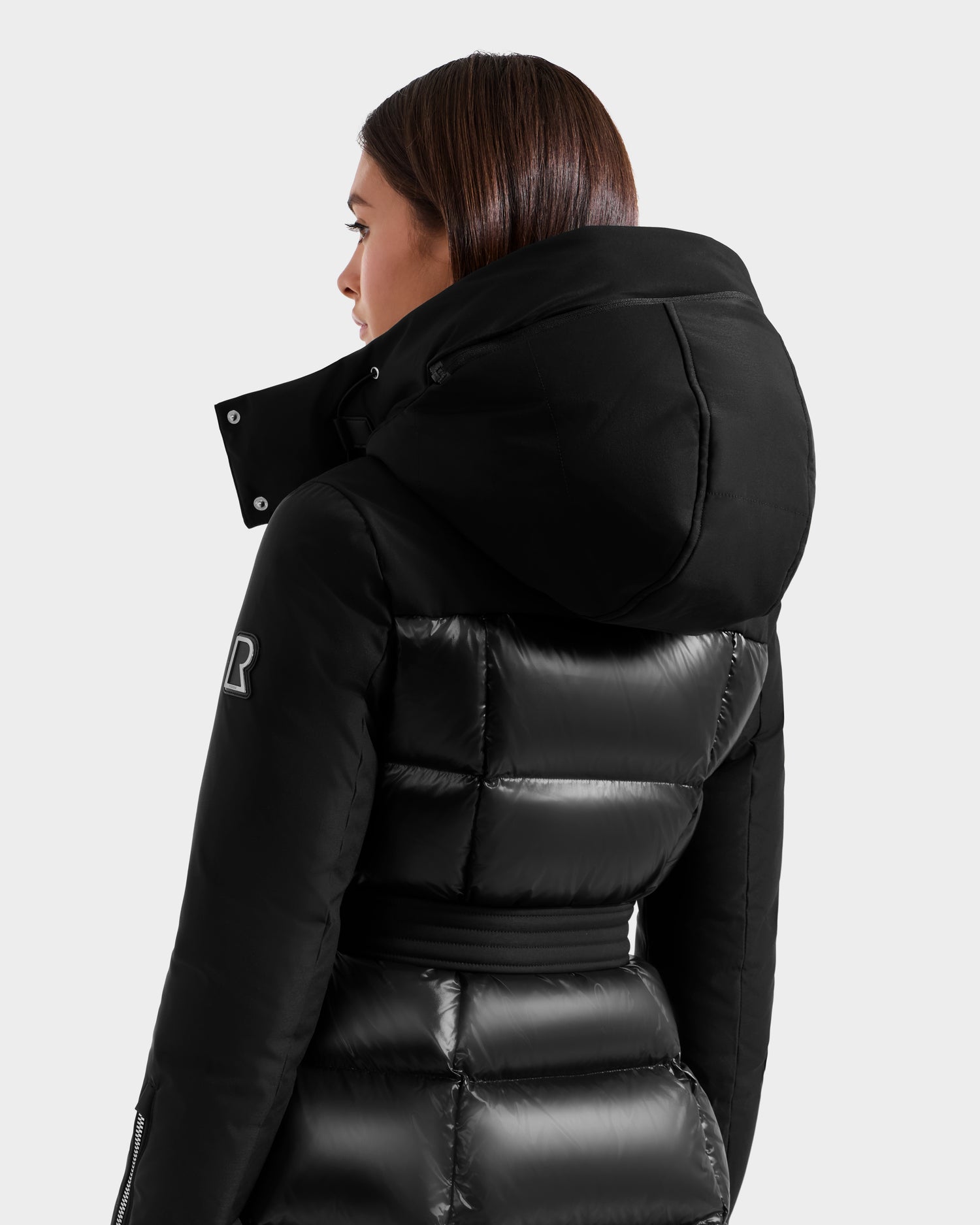 A woman with straight brown hair models the Rudsak CAT H - 8125588 Black women's down puffer coat with a large hood and belt, standing mostly turned away from the camera against a plain light background.