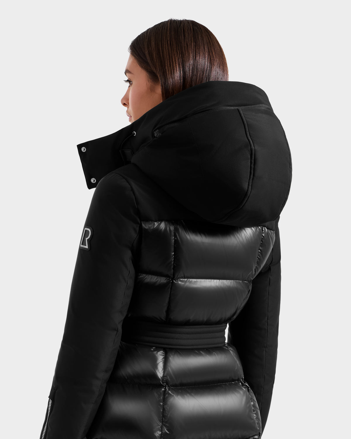 A woman with straight brown hair models the Rudsak CAT H - 8125588 Black women's down puffer coat with a large hood and belt, standing mostly turned away from the camera against a plain light background.