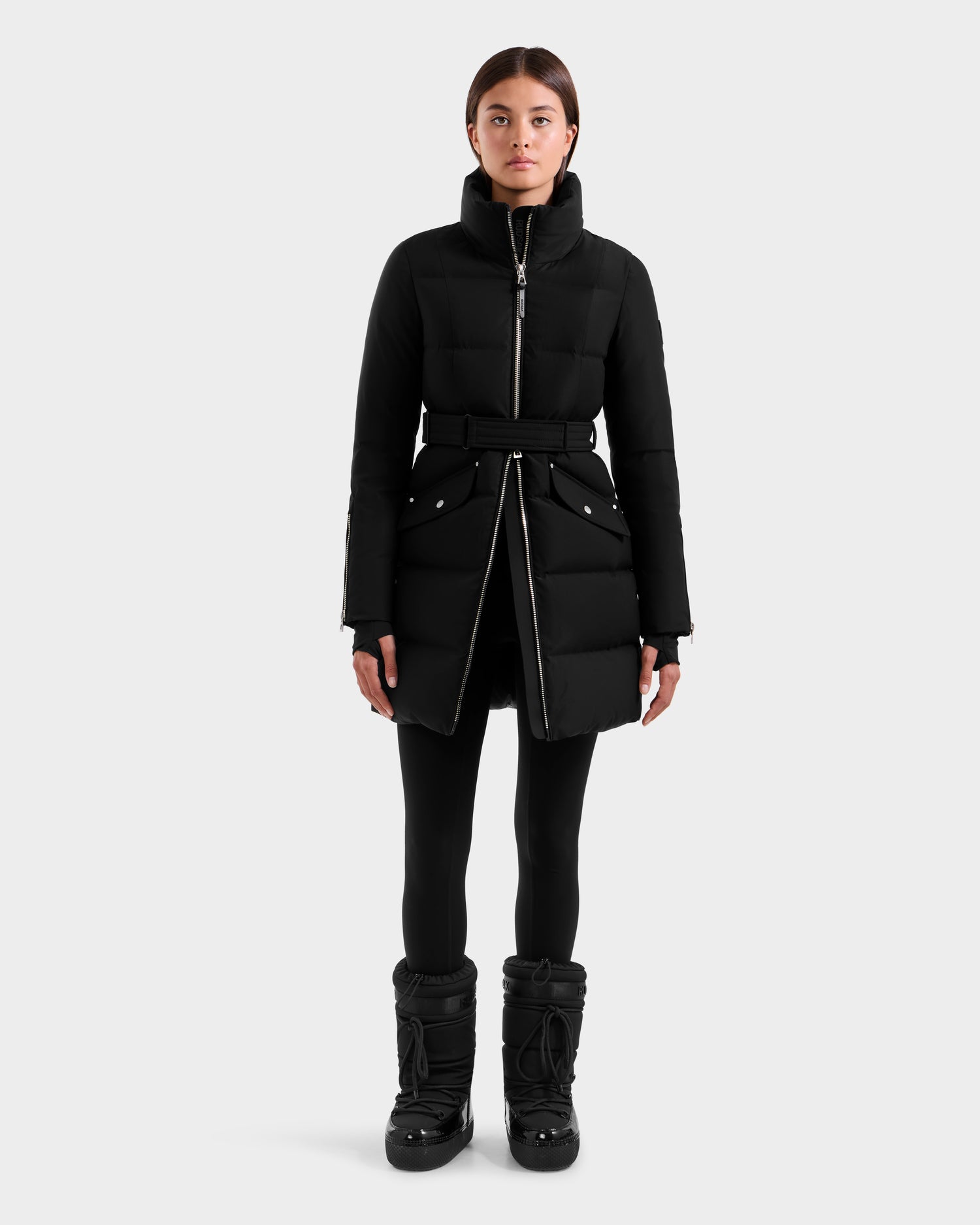 A woman stands facing forward against a light gray background, wearing the Rudsak CAT H - 8125588 Black women’s down puffer coat with a belted waist, paired with black leggings and black snow boots.